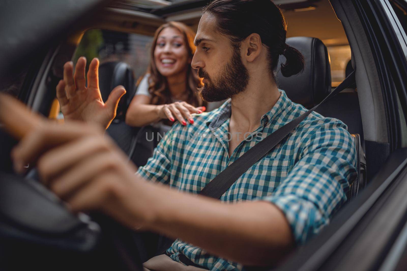 A handsome young man is driving a car and young smiling woman is sitting on the back seat tapping his shoulder and talking something to him.