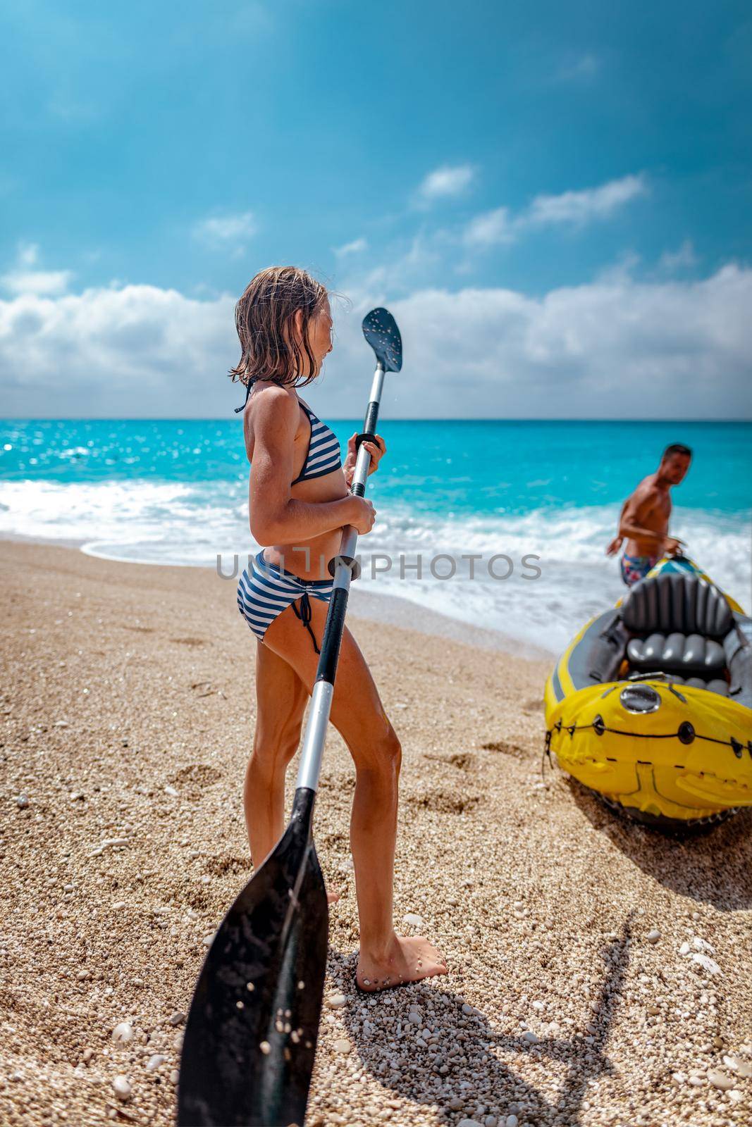 Little girl playing with a paddle beside a kayak which her father prepares for sailing in the sea.
