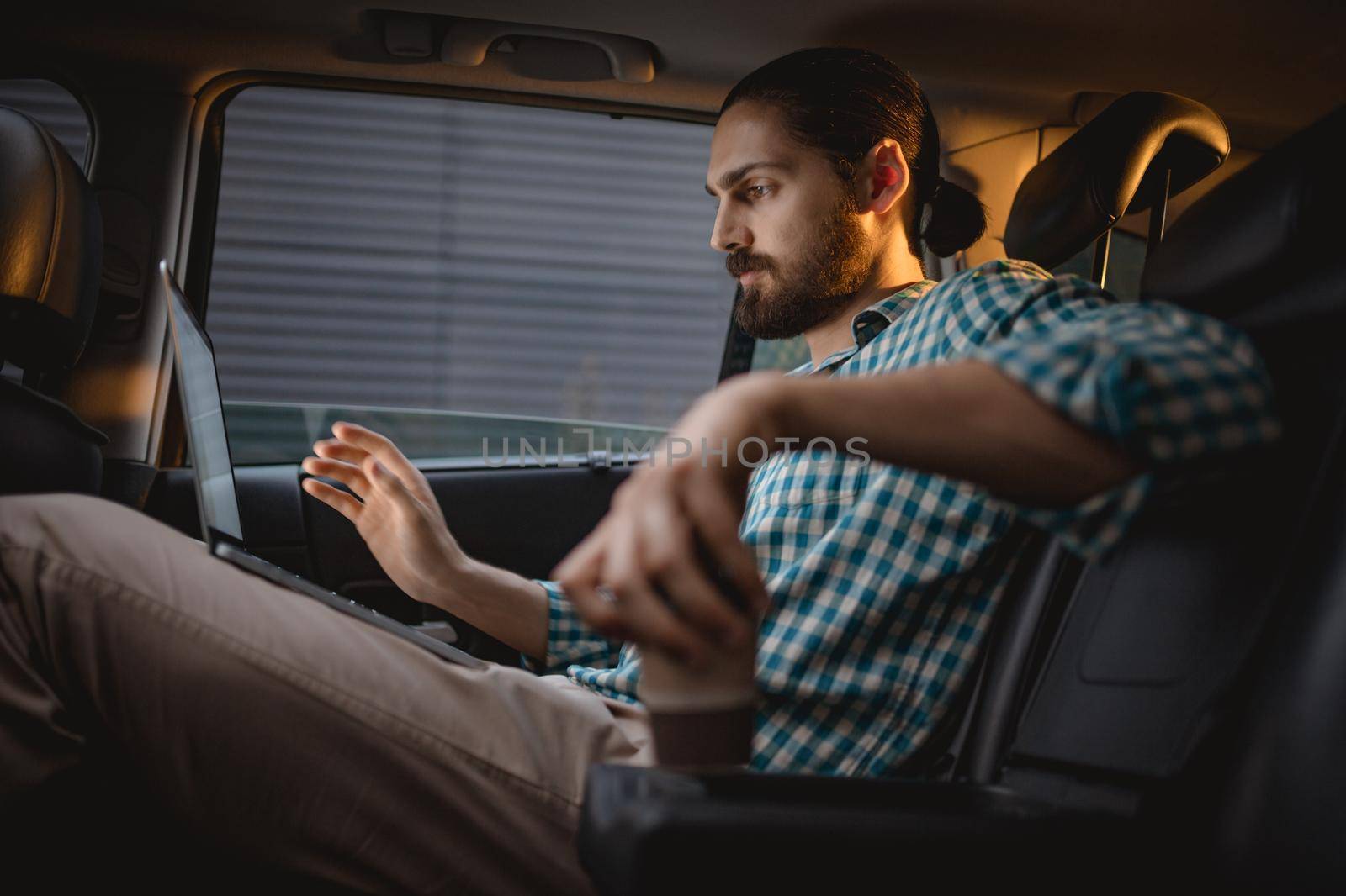 Young confident businessman is looking at laptop on the back seat in car.
