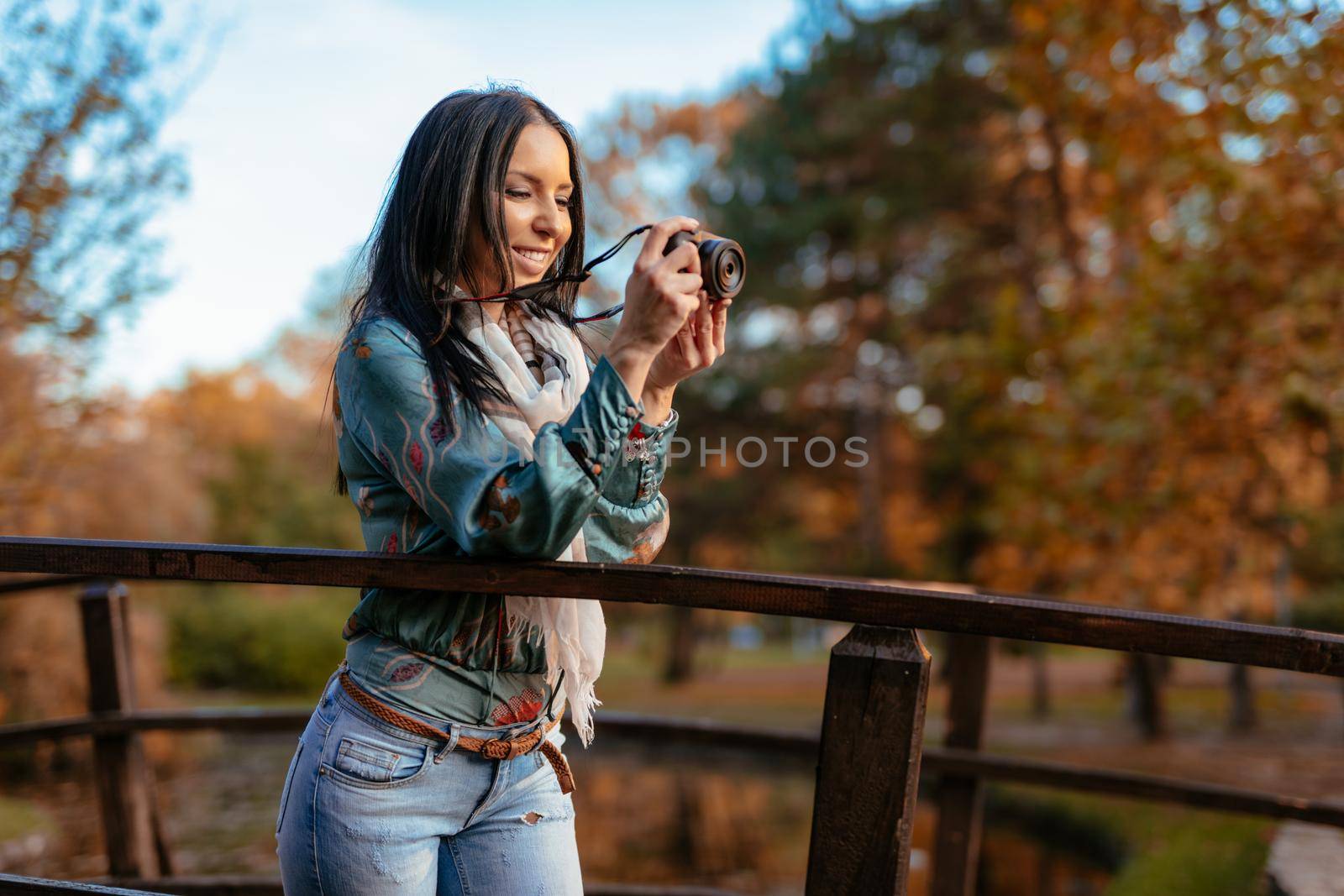 Woman photographer holding professional digital camera and taking photo on the bridge in autumn city park.  