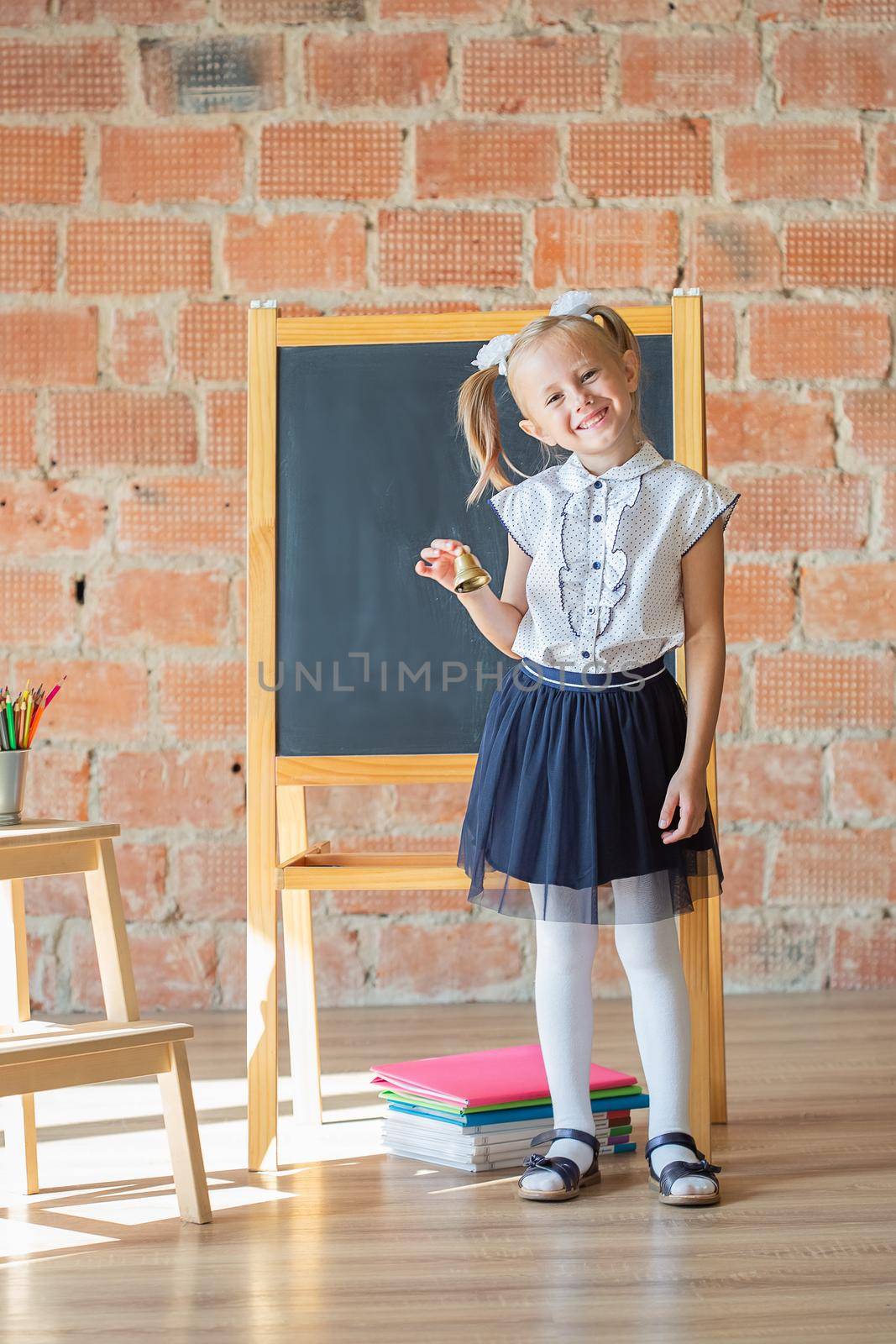 Adorable caucasian private school kindergarten girl smiling in front of blackboard with a bell in her hands, back to school concept