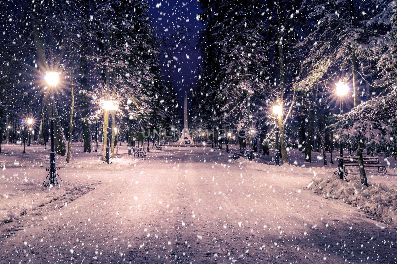Snowfall in a winter park at night with christmas decorations, lights and  pavement covered with snow. Falling snow.