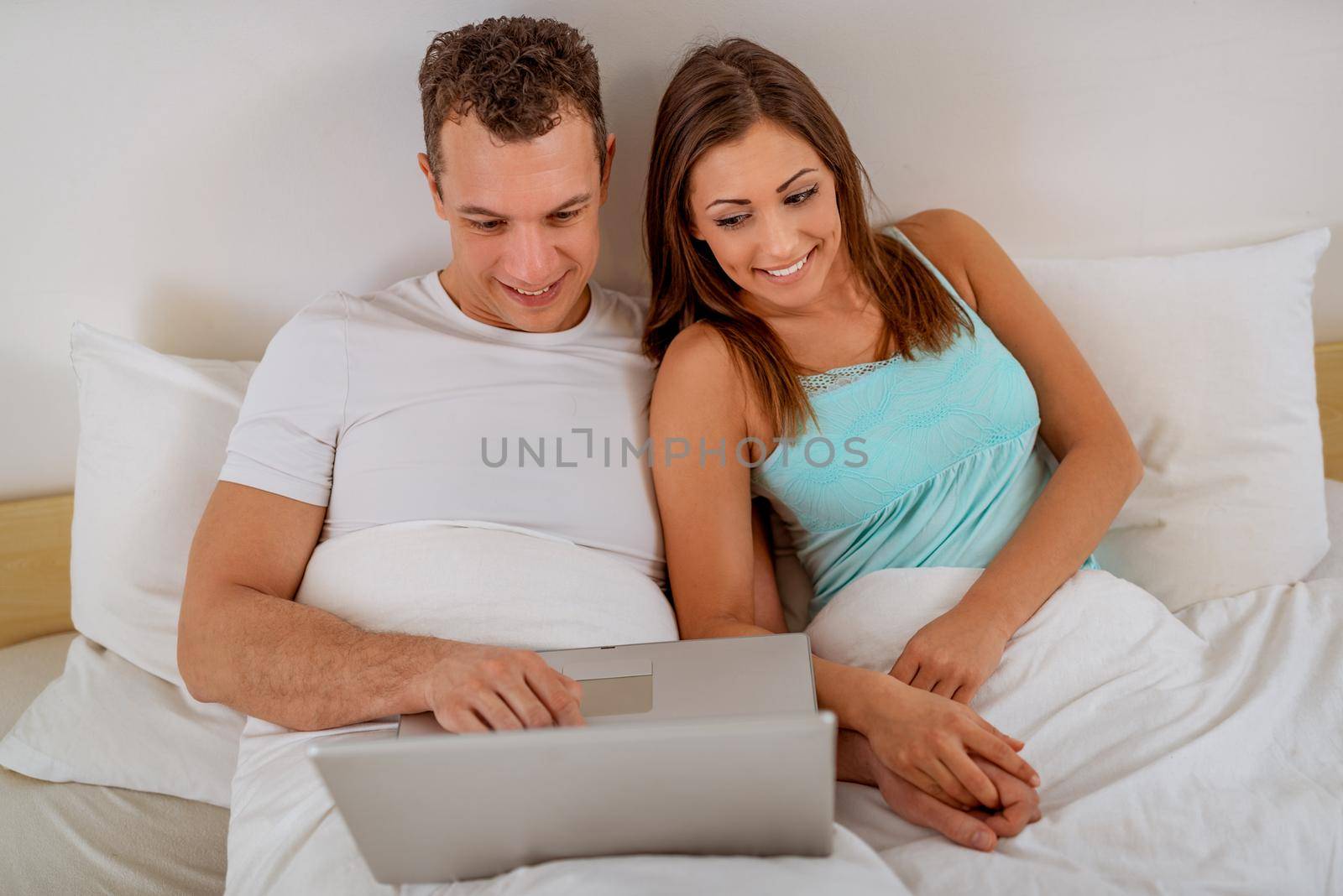 Beautiful young smiling couple enjoying in morning. They is sitting in bed with laptop and surfing on internet.