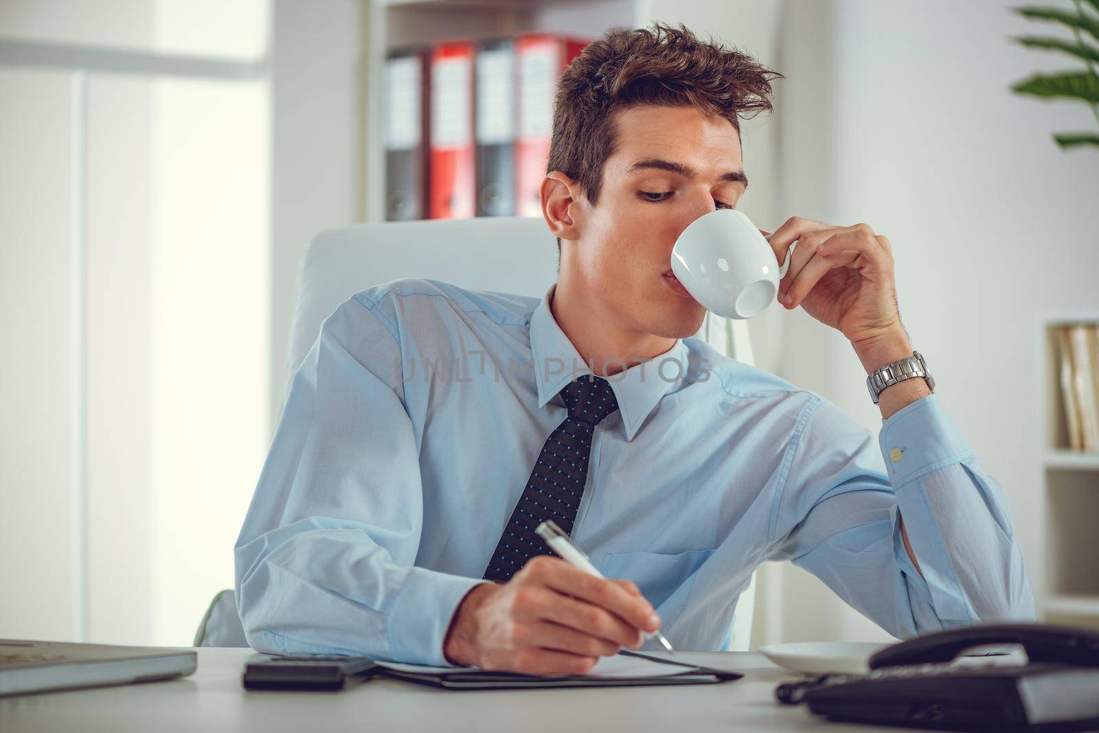 Executive male working at desk looking at documents and accounts. He is drinking coffee, holding a pen and looking concentrated and serious