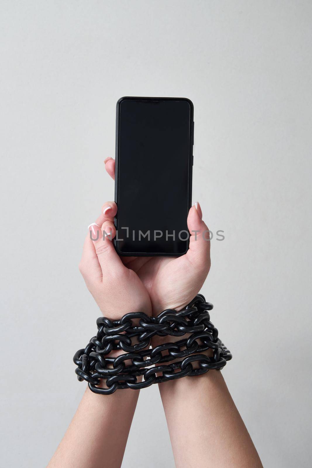 Iron chain that ties together hand and smartphone in concept of social media and internet addiction on gray background by Mariakray