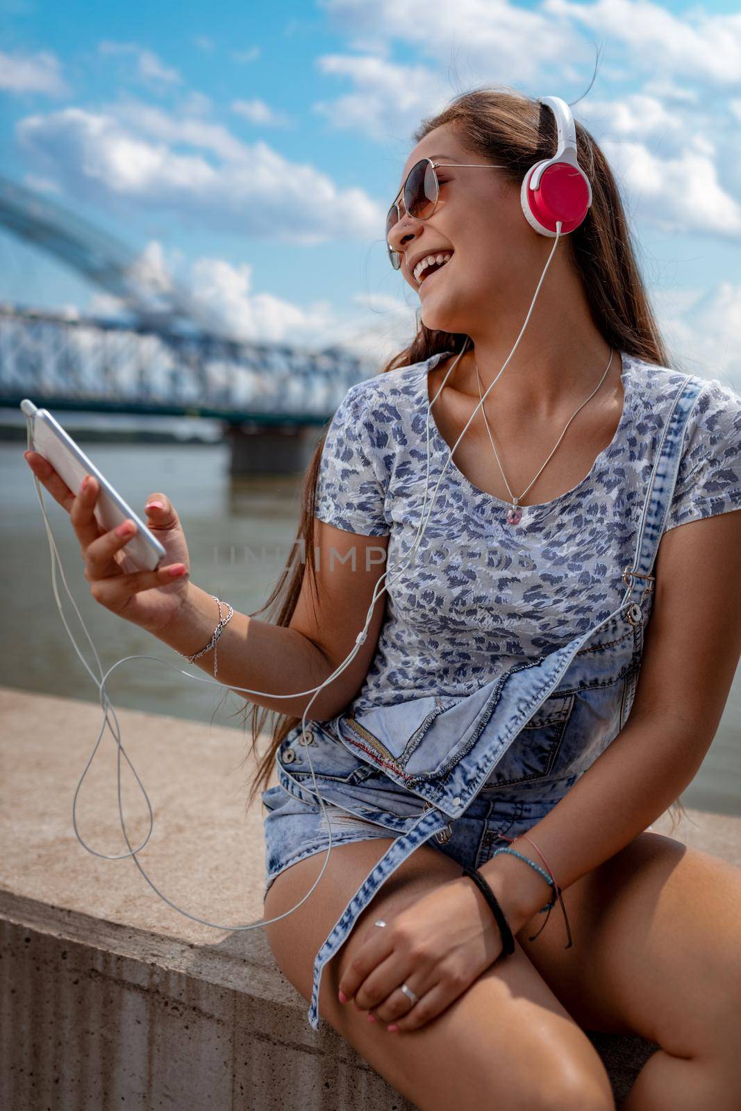 A beautiful smiling girl listening music from your phone and enjoying on the city rivershore on a beautiful summer day. 