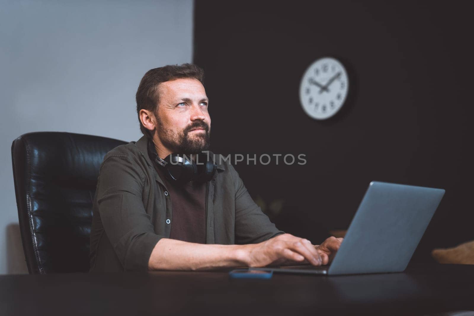 Worker daydreaming while sitting in office. Man dressed in casual wear, typing on laptop. However, distant gaze and relaxed posture suggest that he lost in thoughts, indulging in moment of dreaming. High quality photo