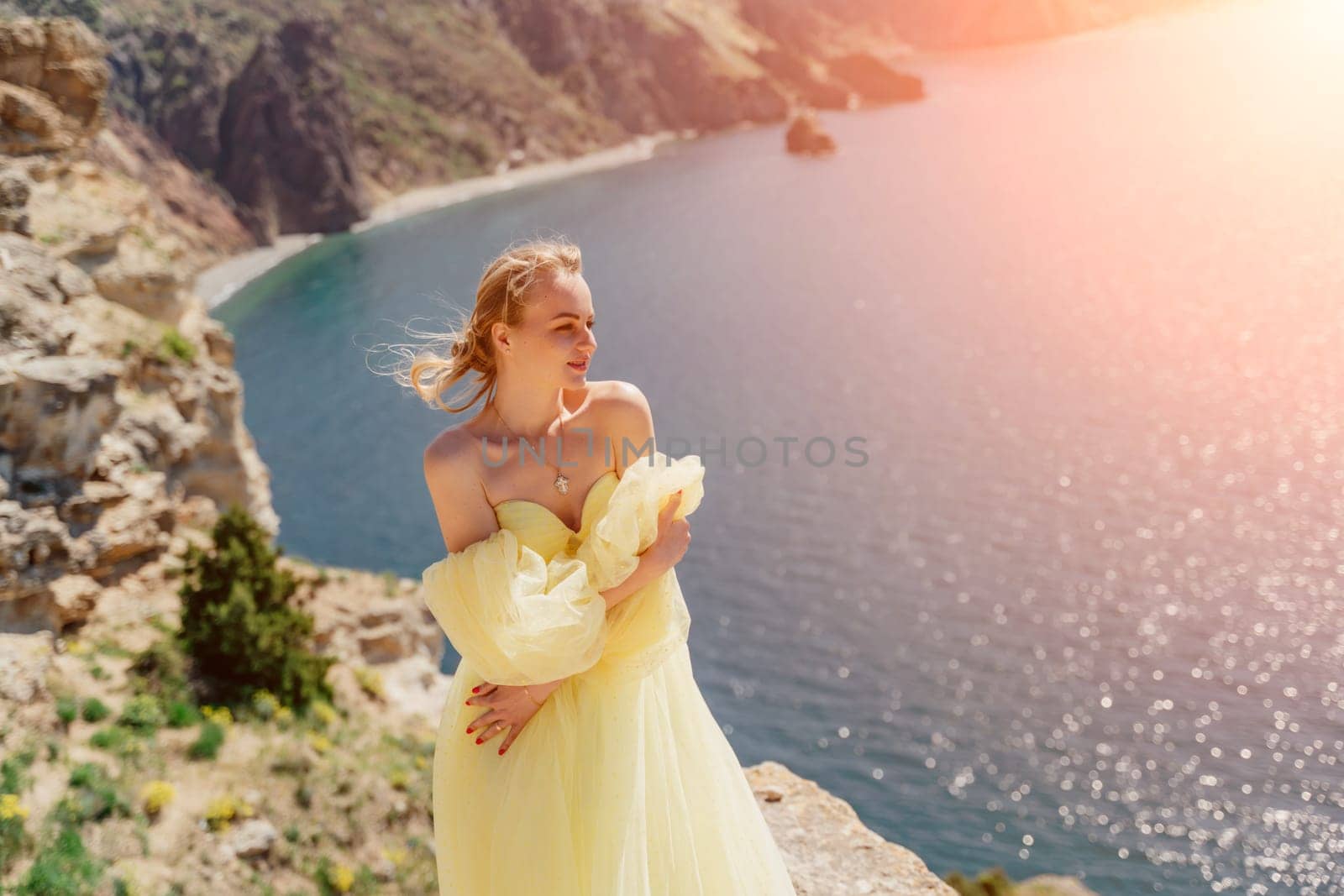 Woman yellow dress sea. Side view Young beautiful sensual woman in yellow long dress posing on a rock high above the sea at sunset. Girl in nature against the blue sky.