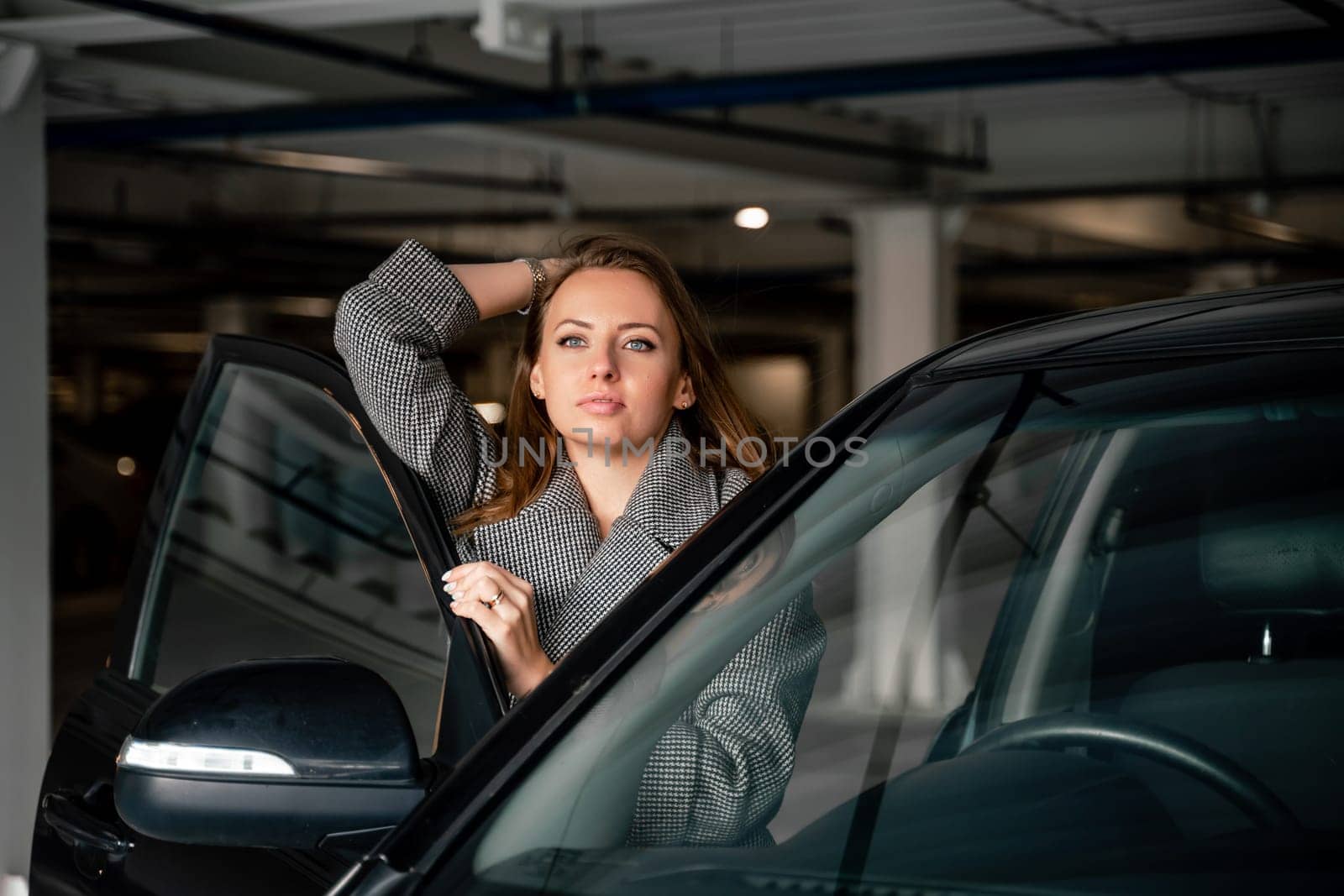 Happy woman car. she stands next to the car in the underground parking. Dressed in a gray coat, holding a glass of coffee in her hands, a black car. by Matiunina