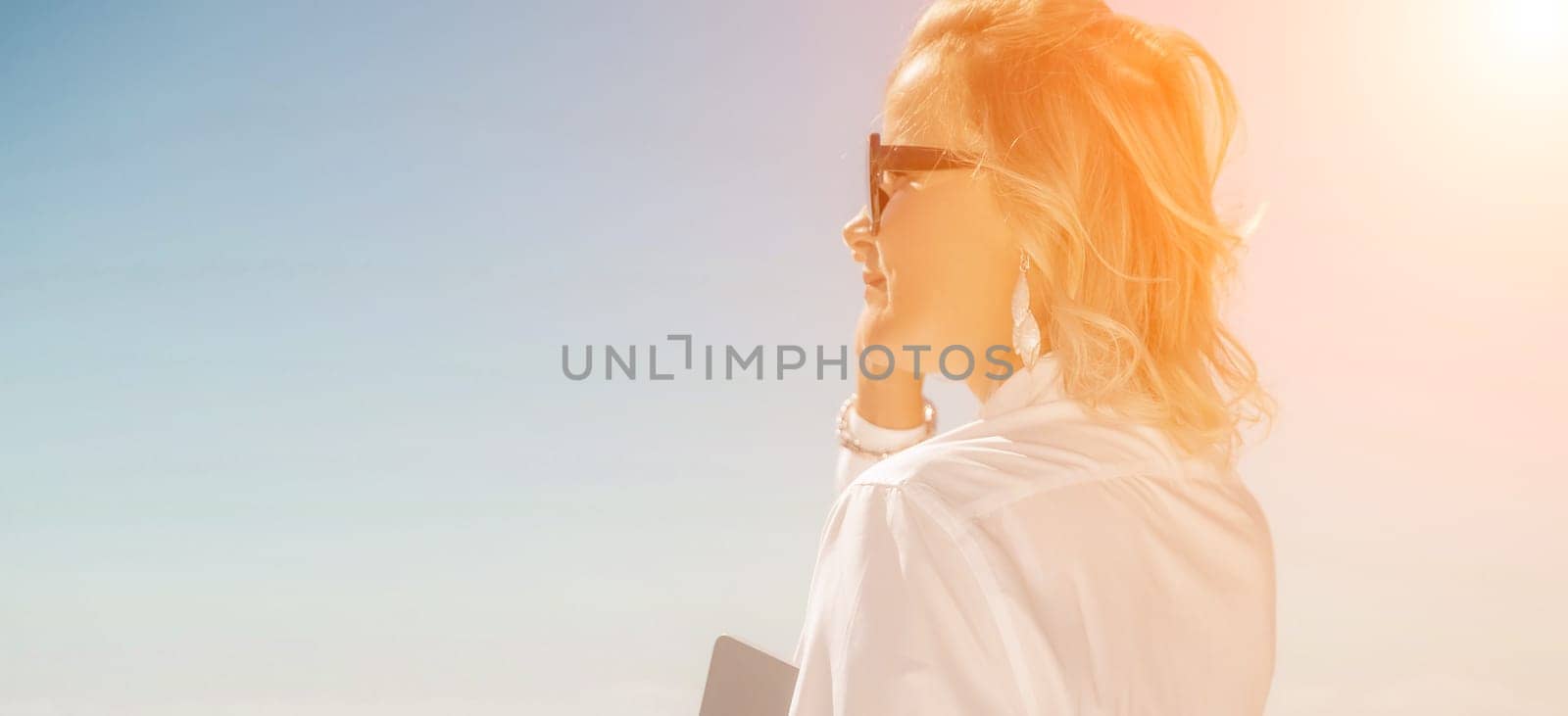 Freelance women. Pretty middle aged woman with computer and phone outdoors with beautiful sea view. The concept of remote work. by Matiunina