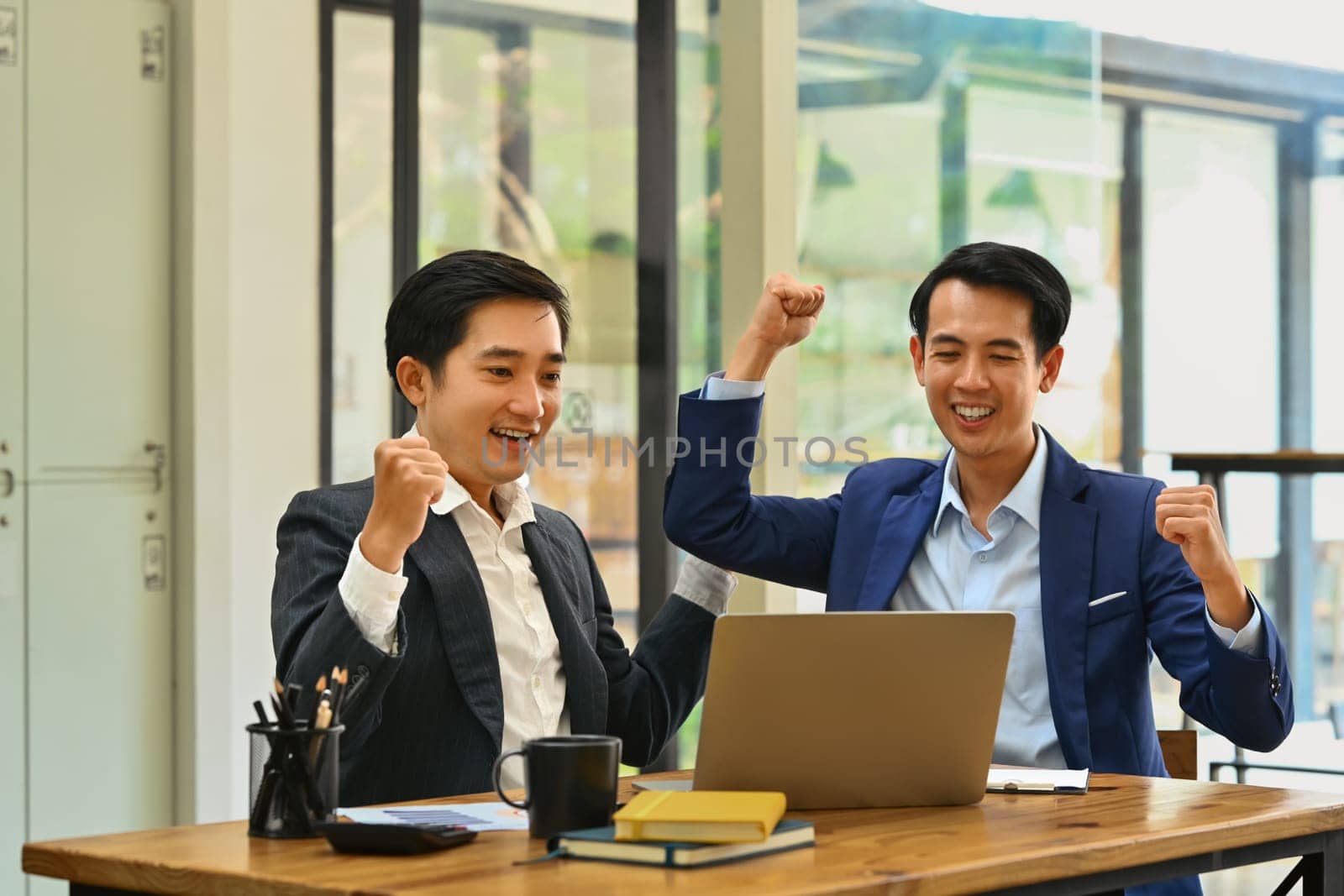 Excited business people looking at laptop screen excited by good news online, celebrating success raising hands up.