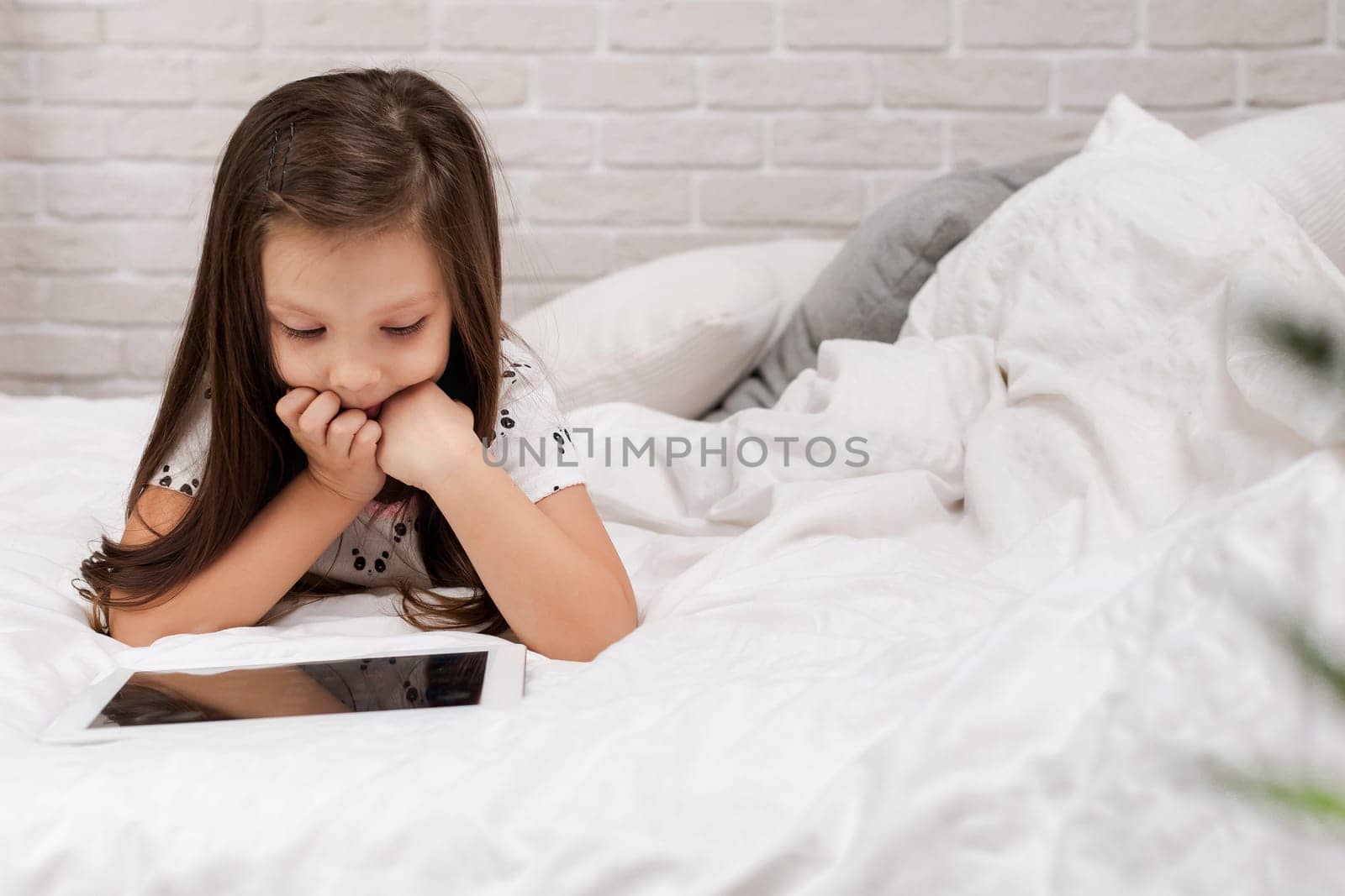 cute little child girl lies in bed uses digital tablet. child playing on tablet pc.