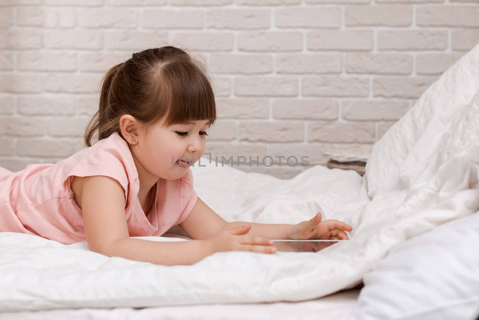 cute little child girl lies in bed uses digital tablet. child playing on tablet pc. space for text