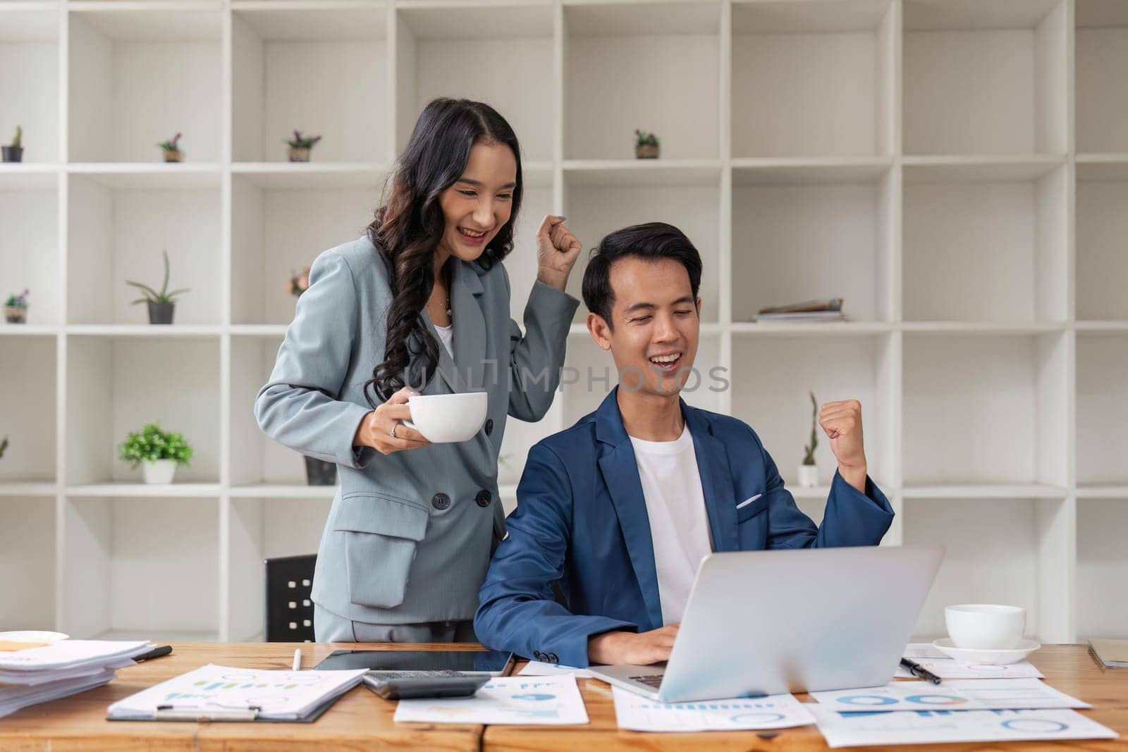 Two asian business people successful excited raised hands rejoicing with a laptop computer in office.