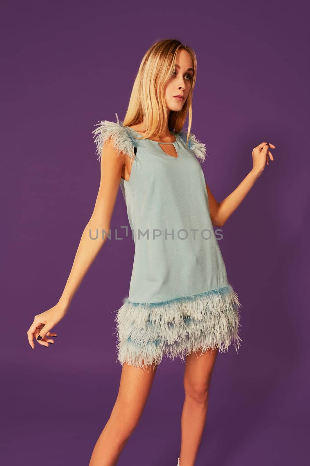 Young attractive woman with blonde long hair, dressed in elegant cocktail dress with feathers, posing at studio before camera