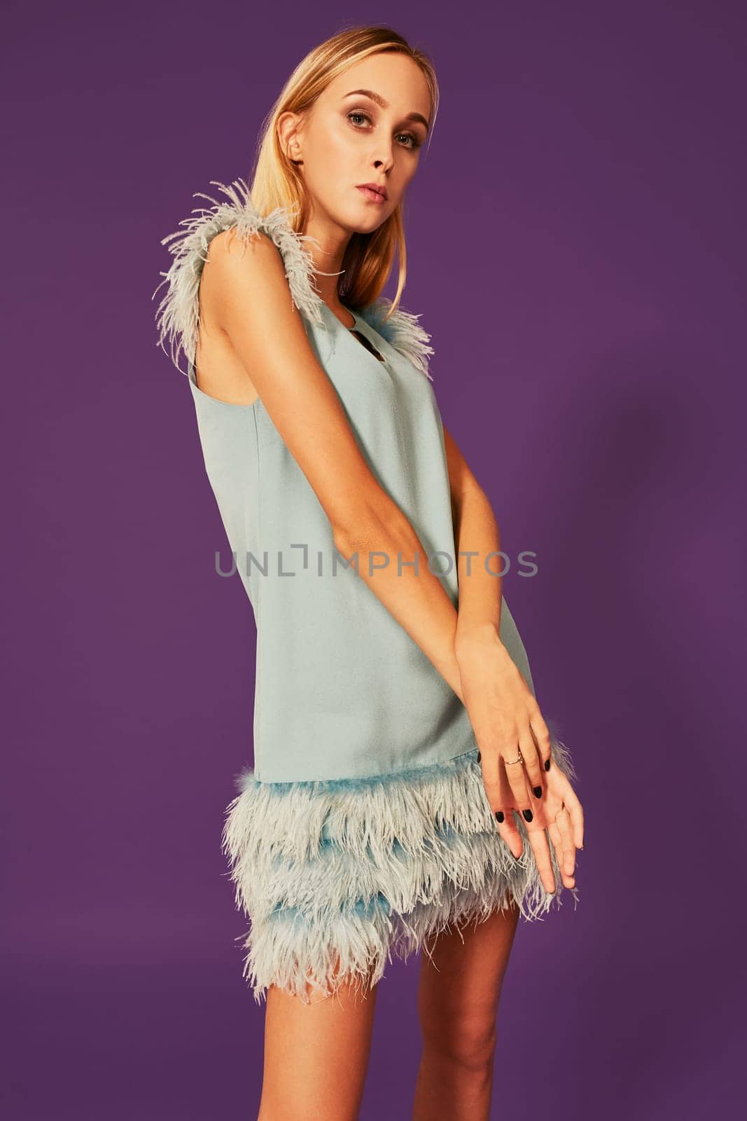 Young attractive woman with blonde long hair, dressed in elegant cocktail dress with feathers, posing at studio before camera with crossed hands