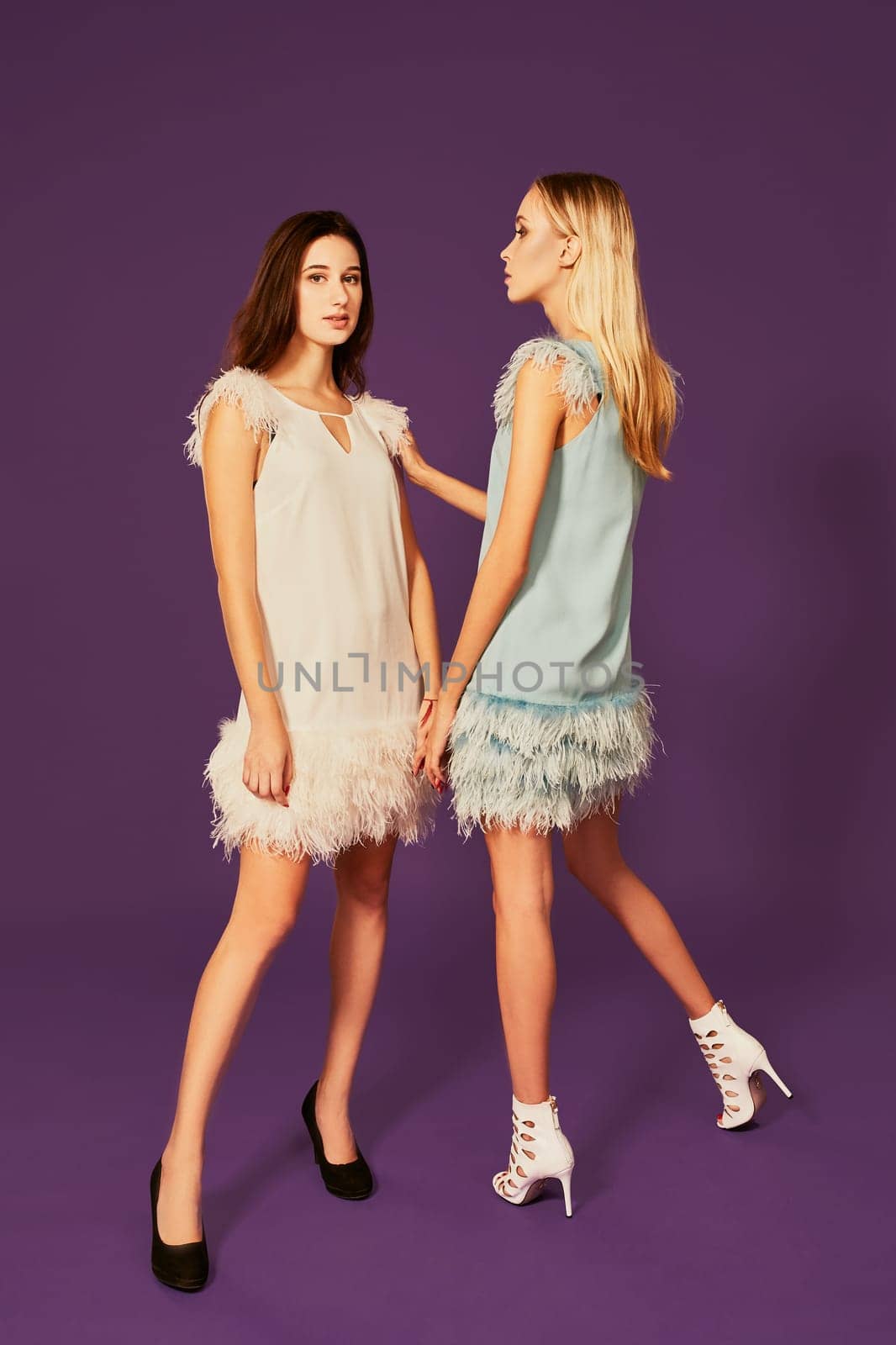 Studio fashion portrait of two elegant young women in cocktail light dresses posing in studio. Blonde and brunette hold hands, Black and white shoes