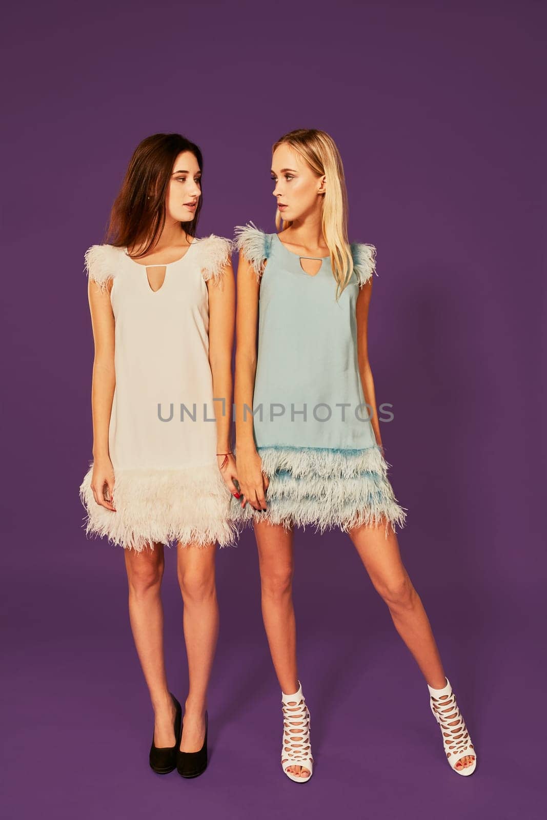Studio fashion portrait of two elegant young women in cocktail light dresses posing in studio. Blonde and brunette hold hands, Black and white shoes