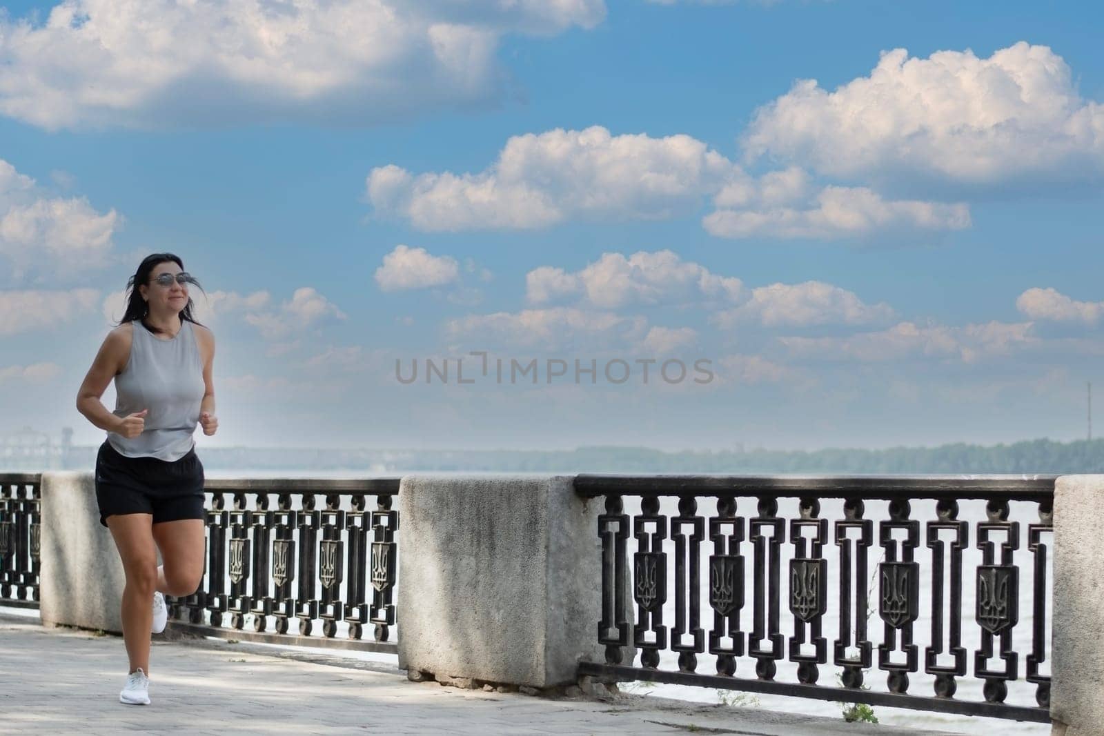 Sports concept. A beautiful, tall, athletic girl in short shorts runs along the embankment near the river, is engaged in active jogging outdoors in the summer. Outdoor fitness.