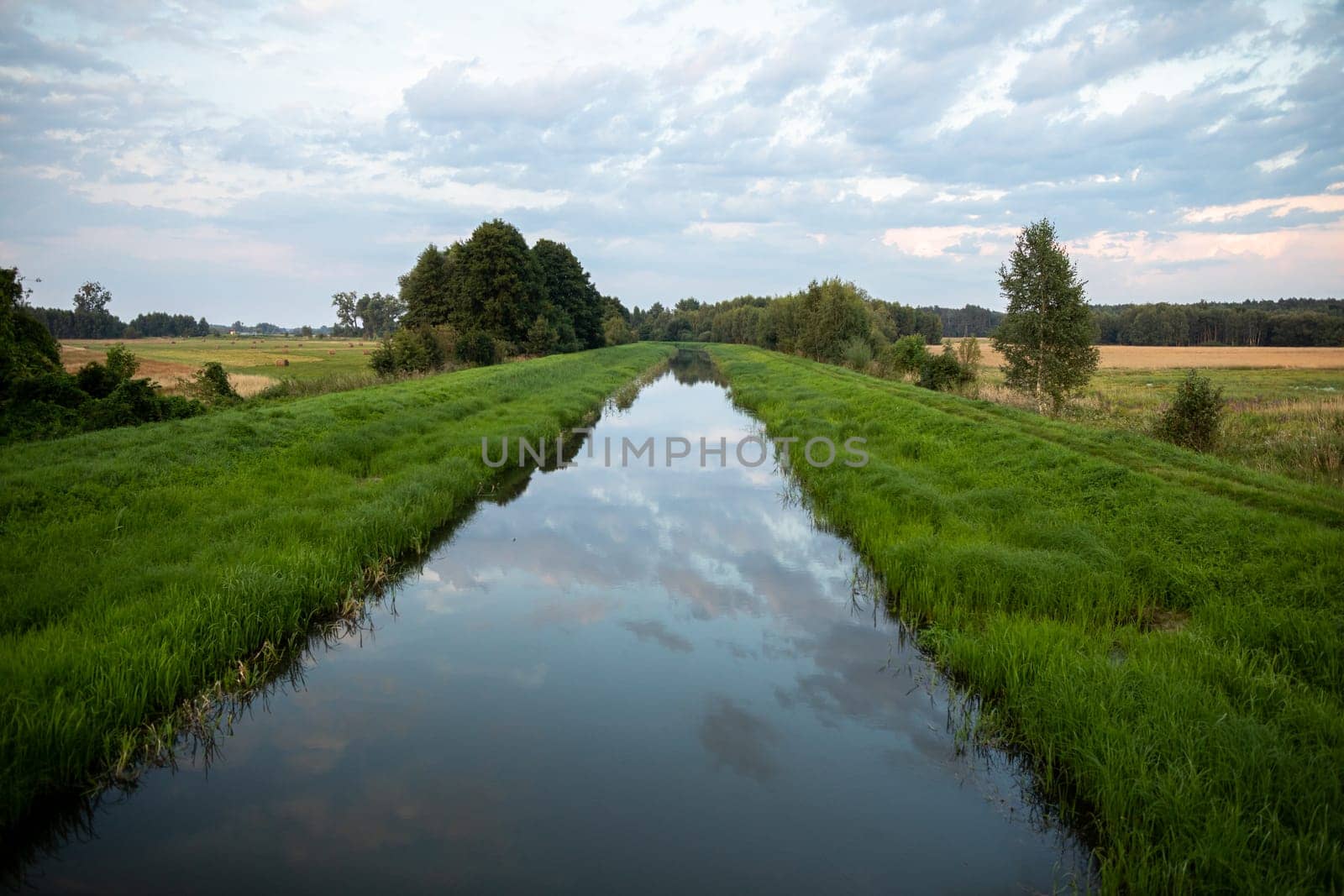 With a slow current, the river flows straight amidst green grasses and agricultural fields. The overcast sky is clearly reflected in the calm river.