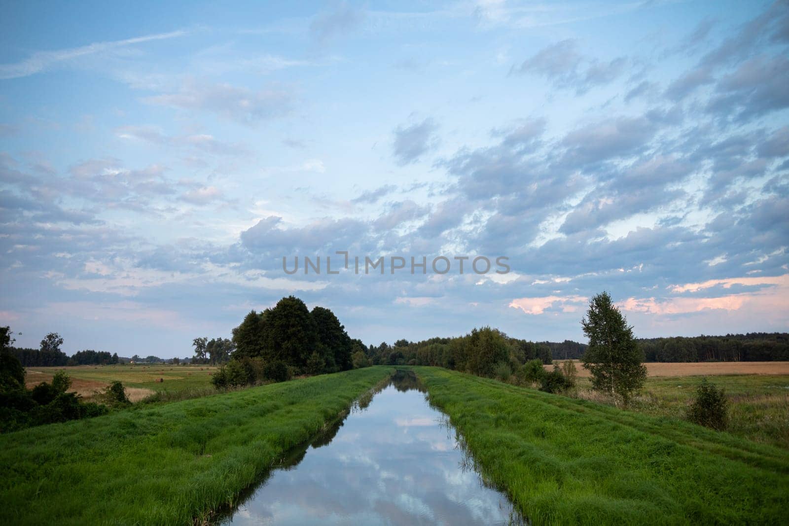 With a slow current, the river flows straight amidst green grasses and agricultural fields. The overcast sky is clearly reflected in the calm river.