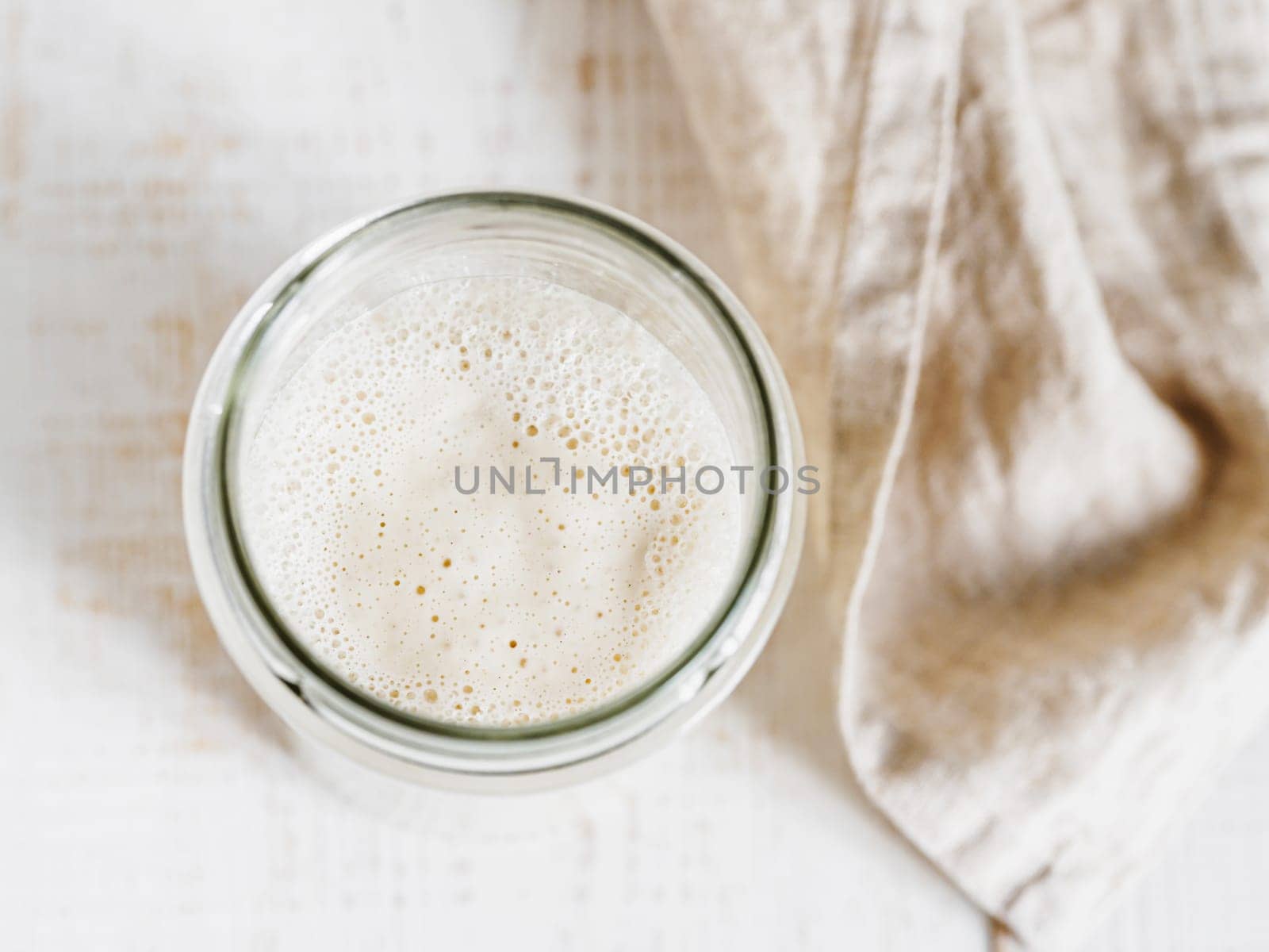Wheat sourdough starter. Top view of glass jar with sourdough starter on white wooden background. Copy space for text or design.