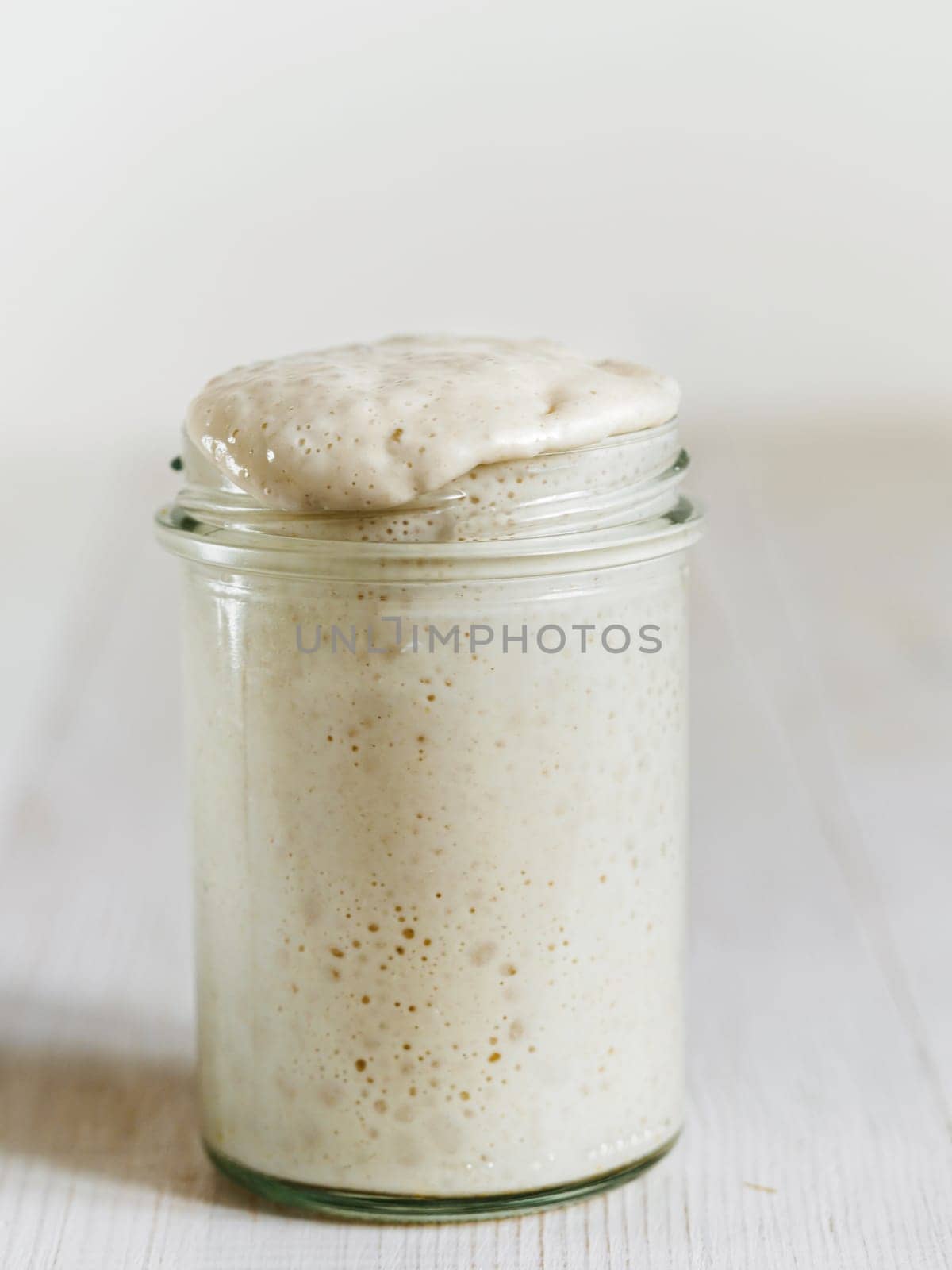 Wheat sourdough starter. Glass jar with sourdough starter on white wooden tabletop. Copy space for text or design. Vertical.