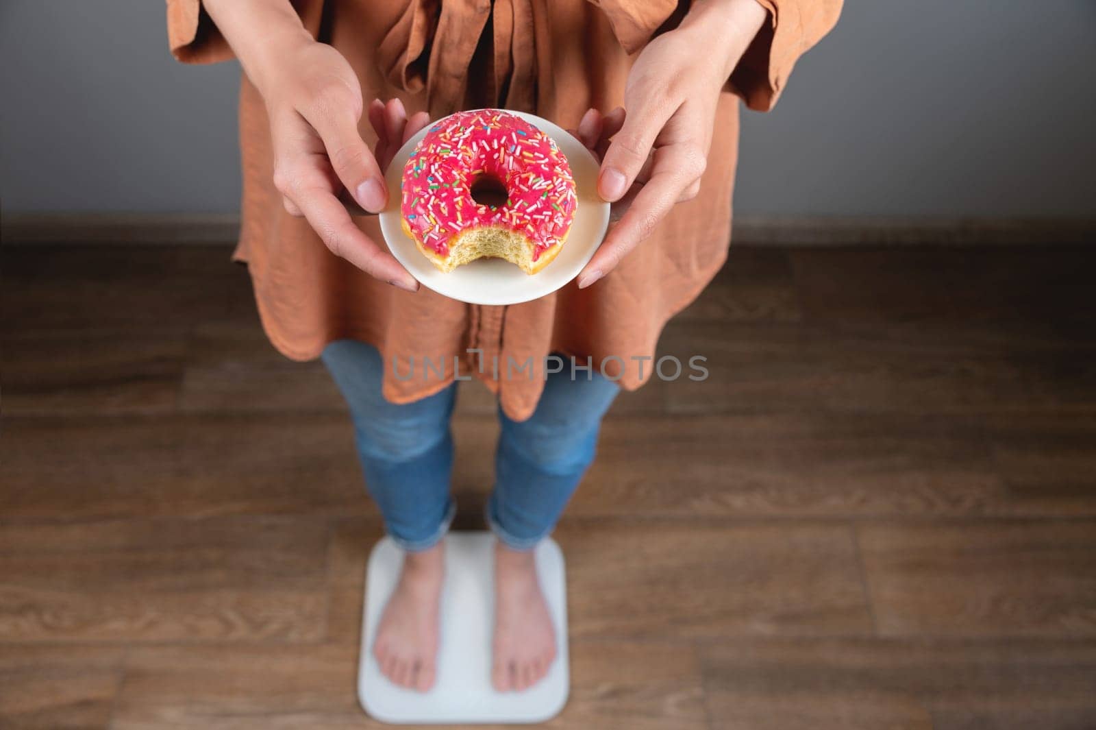 Diet. A woman measures body weight on a scale, holding a donut on a plate. Sweets, unhealthy junk food. Diet, healthy eating, lifestyle. Weight loss. Obesity. View from above.