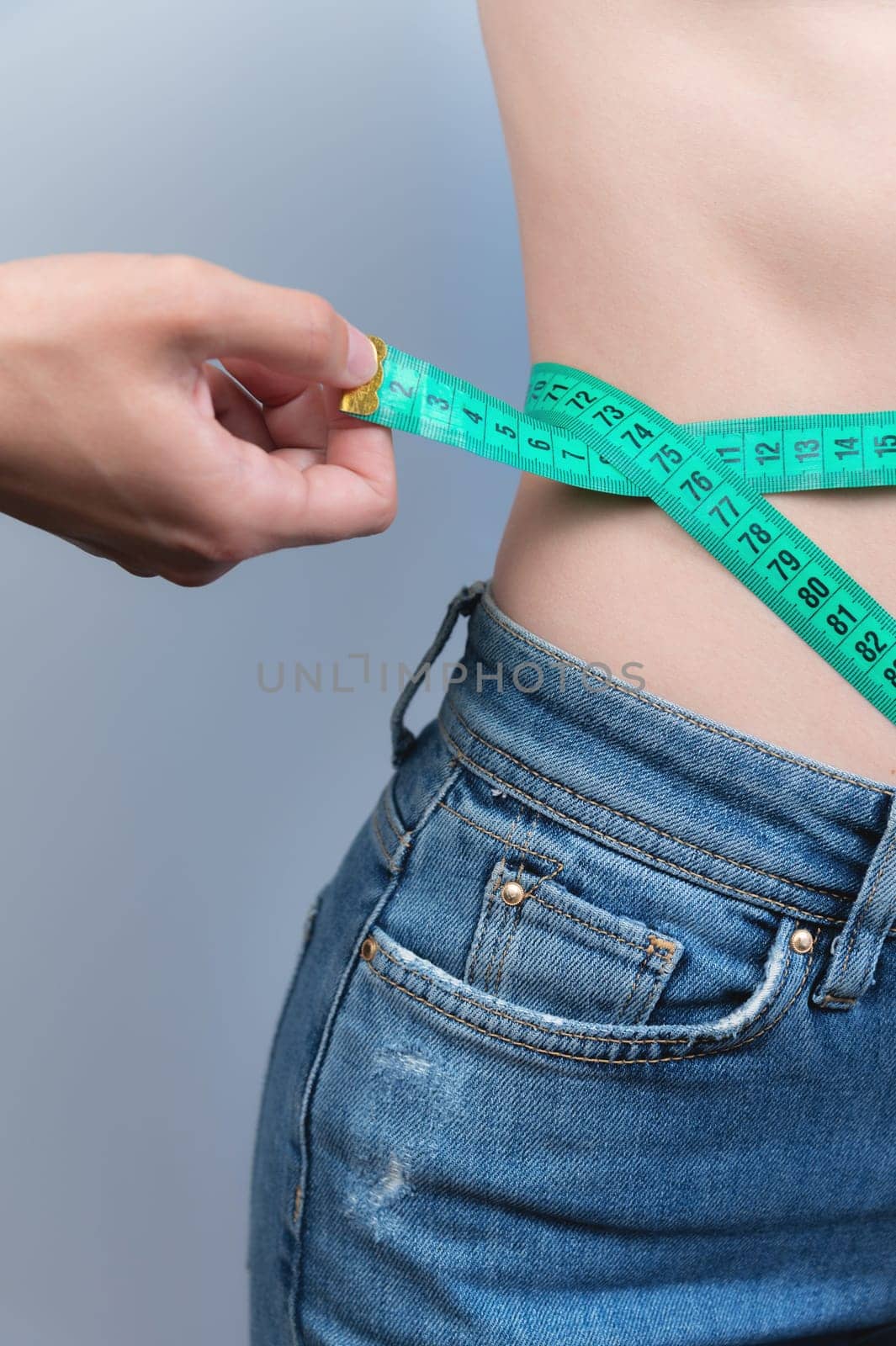 A young woman in denim pants measures her waist with a tape on a gray background. The concept of caring for yourself and your weight.