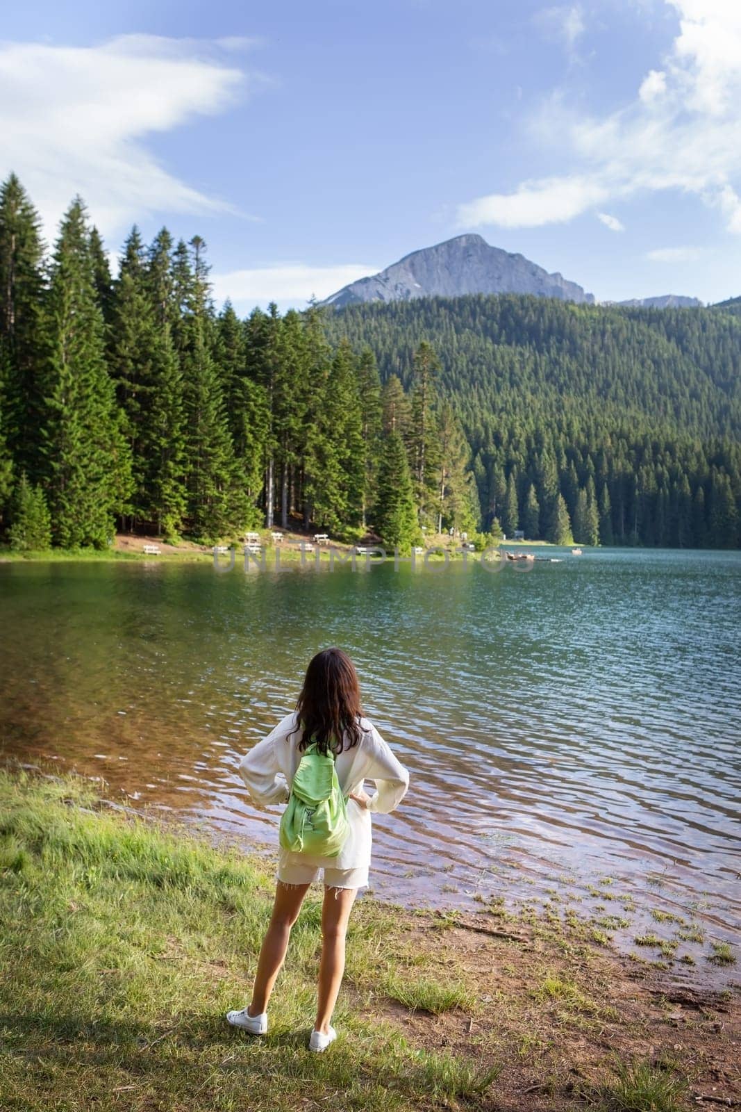 A girl with a bright backpack stands with her back near a mountain lake in the background. Positive young woman traveling on a blue lake outdoors. The girl admires the view. by sfinks