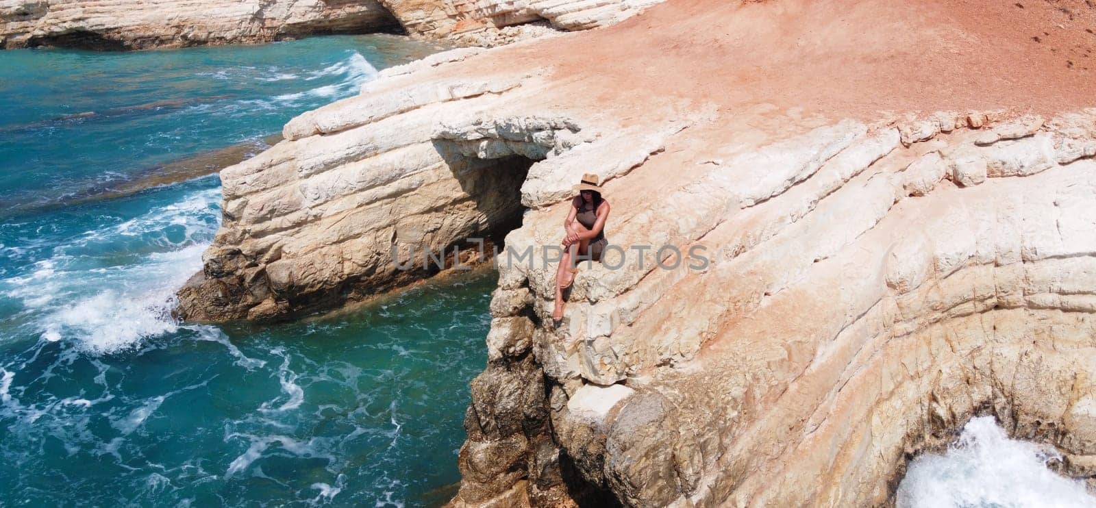 Beautiful girl sitting on a high rock and looking out to sea. top view. Girl in Hat on the edge of cliff. blue sea and high cliffs. Seascape. Girl at sunset. Sea tour. Blue sea, rocks. Cyprus