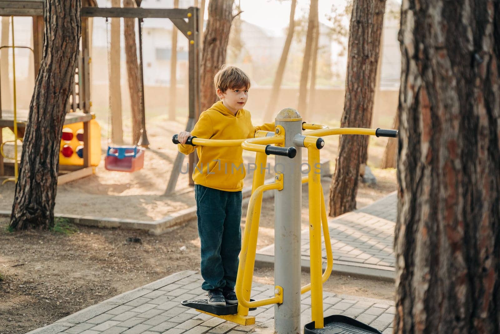 Elementary school boy using fitness exercise equipment in the public city park. Kid playing standing doing exercises on workout equipment in the forest park.