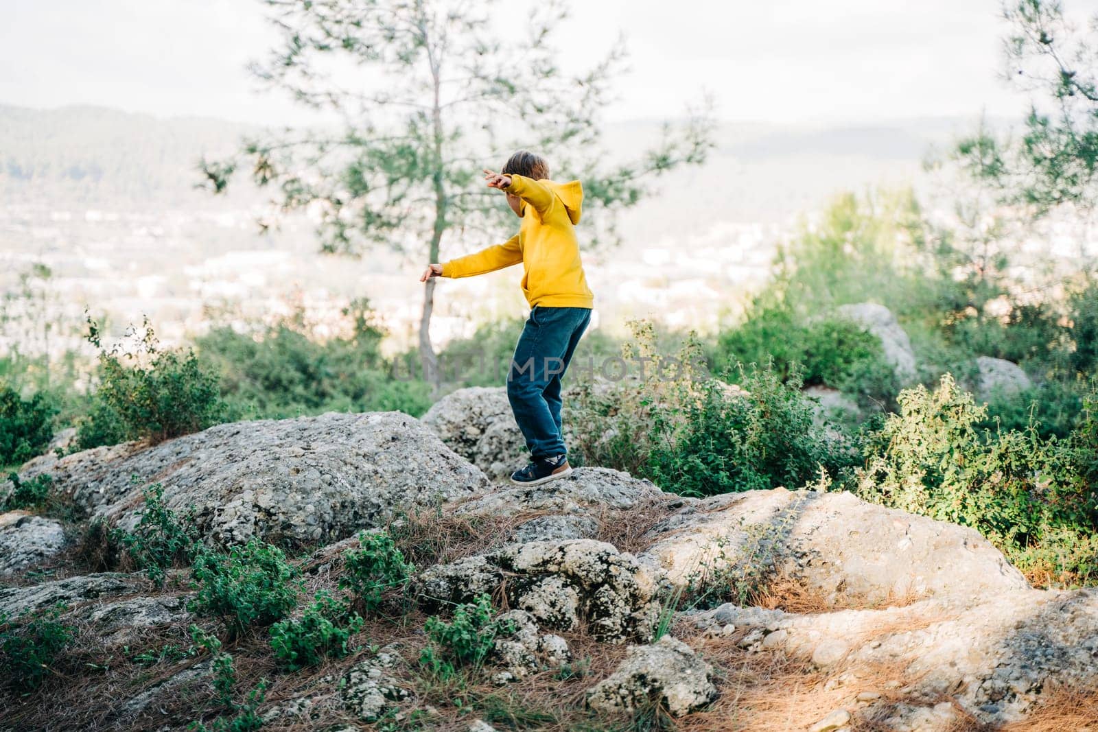 School kid child in vibrant clothes jumping over granite rock boulders in the city park with mountain forest view in the background. Boy leaping across rocks in the park.