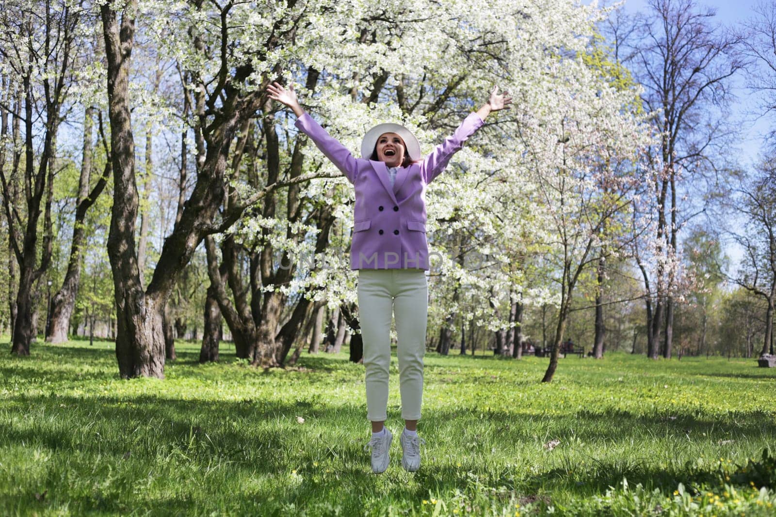 Emotional, joyful bouncing woman in white pants, lilac jacket, and white hat bouncing hard against a blooming tree in a spring park.