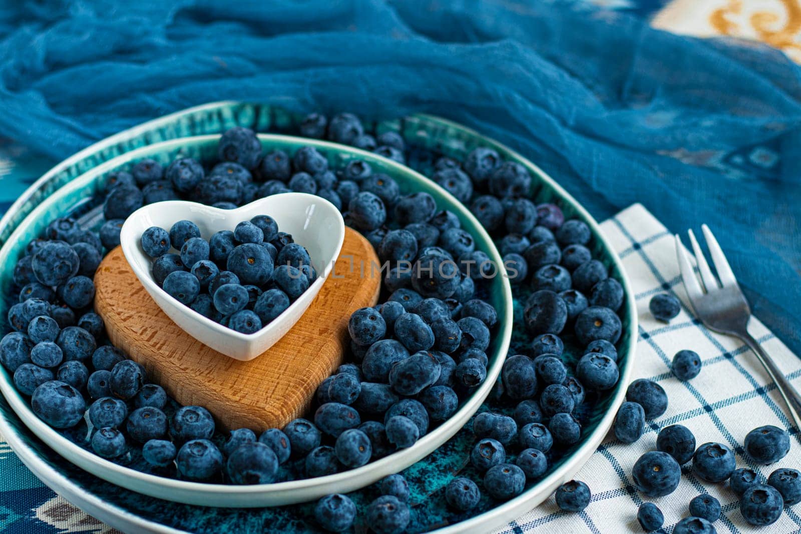 Fresh bilberries on top in heart shaped plate on bilberries background. A lot of fresh bilberries in a blue bowl on bilberries background. High quality photo