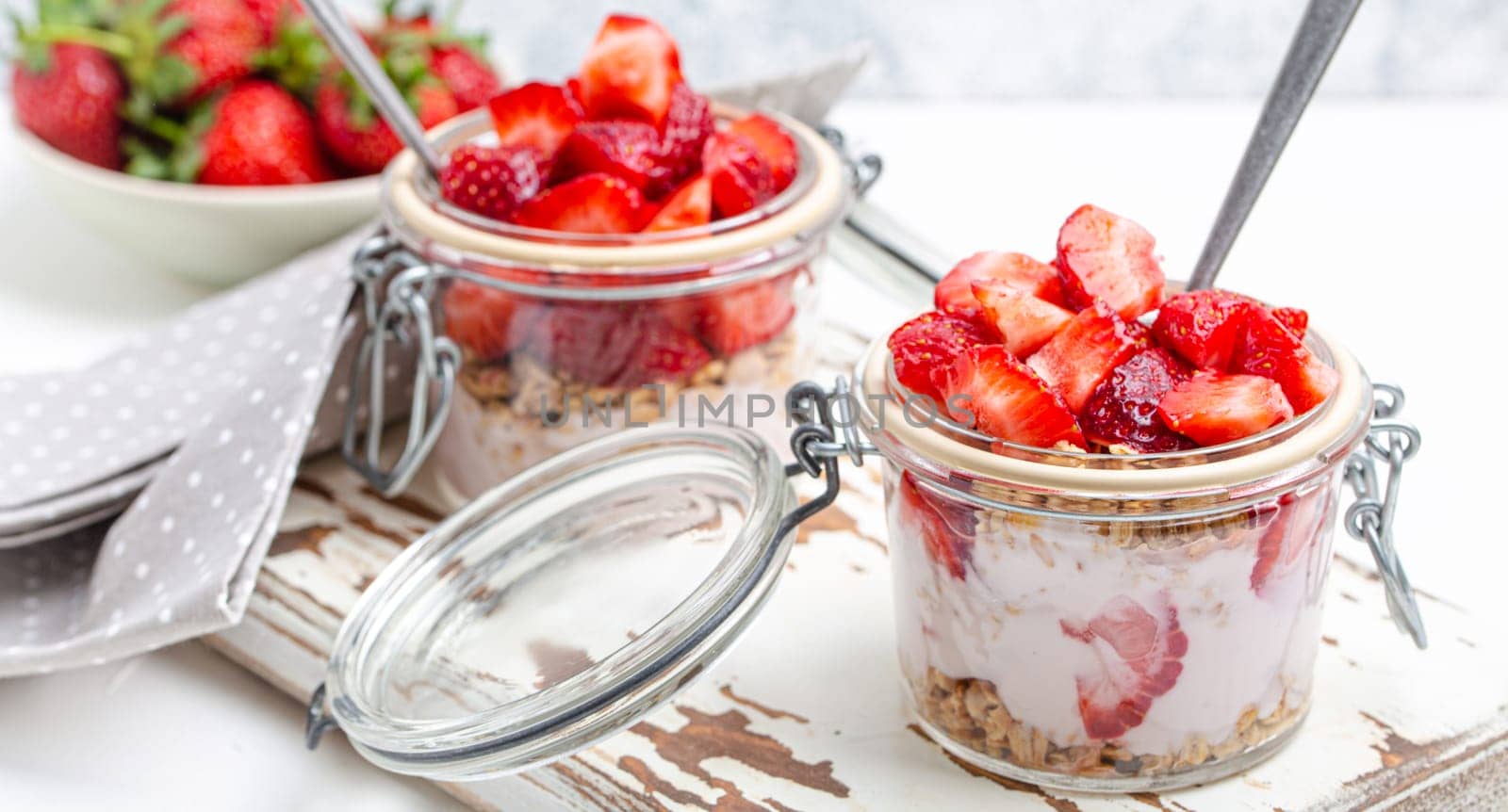 Parfait with Fresh Strawberries, Yoghurt and Crunchy Granola in Transparent Glass Mason Jars on White Rustic Wooden Background from Angle View, Healthy Breakfast or Light Summer Fruit Dessert