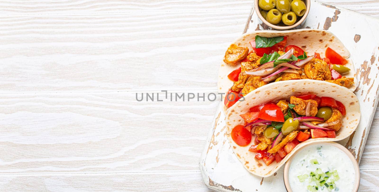 Traditional Greek Dish Gyros: Pita bread Wraps with vegetables, meat, herbs, olives on rustic wooden cutting board with Tzatziki sauce, olive oil top view on white wooden background, space for text.