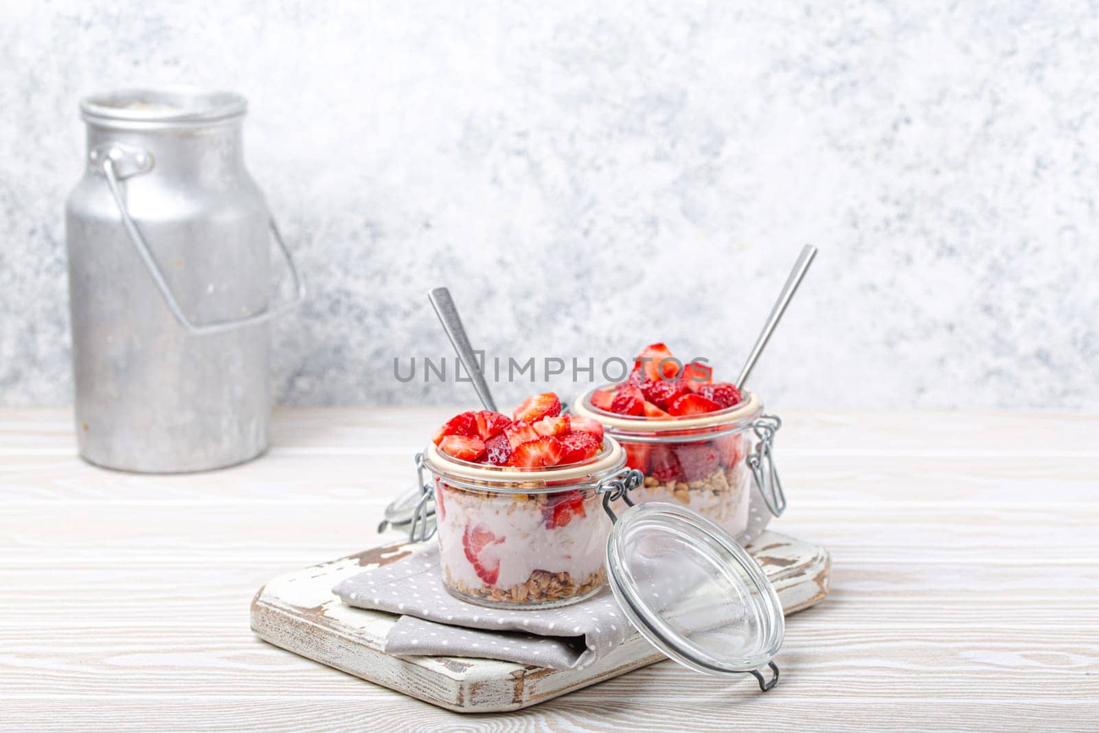 Parfait with Strawberries, Yoghurt and Granola in Transparent Glass Mason Jars on White Rustic Wooden Background Angle View and Aluminium Milk Can, Healthy Breakfast or Summer Dessert, Copy Space by its_al_dente