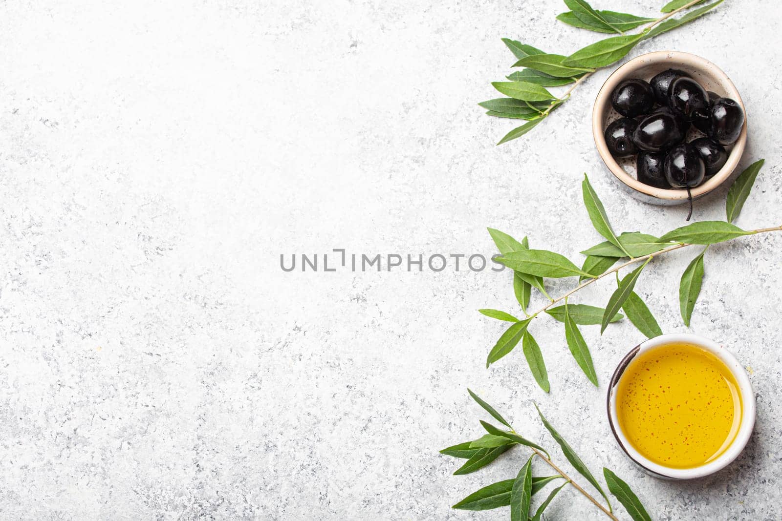 Black olives and extra virgin olive oil in little bowl with olive tree branches top view on white concrete stone background, space for text by its_al_dente