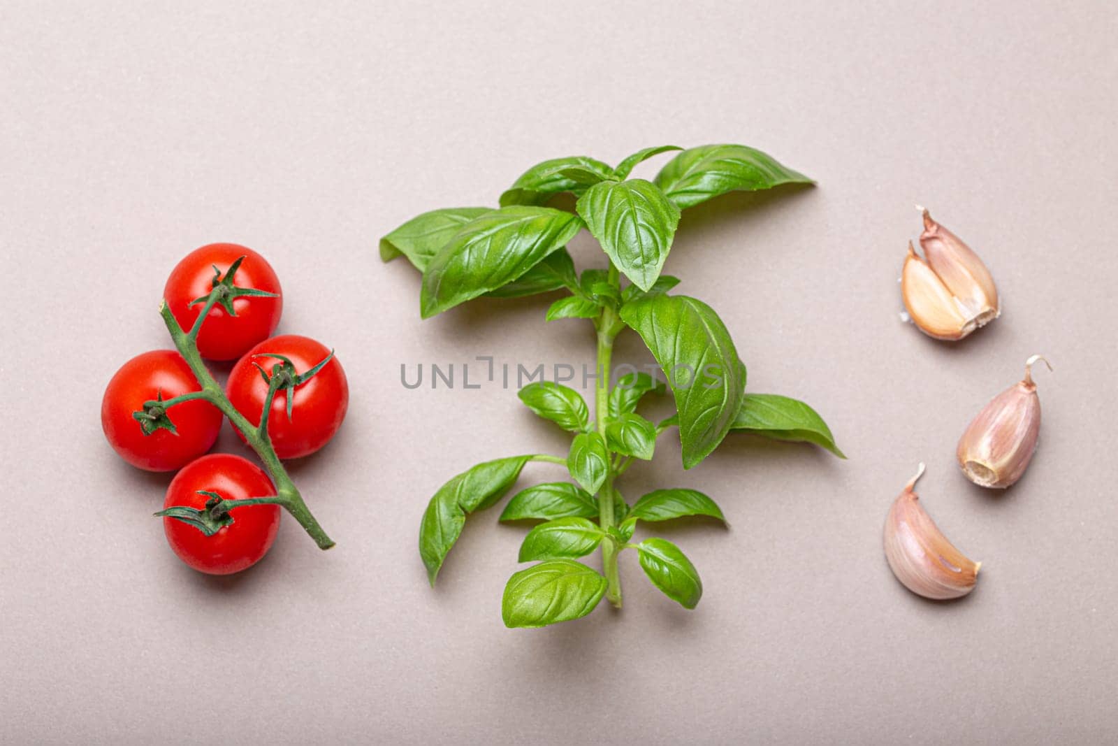 Branch of fresh cherry tomatoes, basil branch and garlic cloves on minimalistic gray clean background, overhead shot. Food ingredients