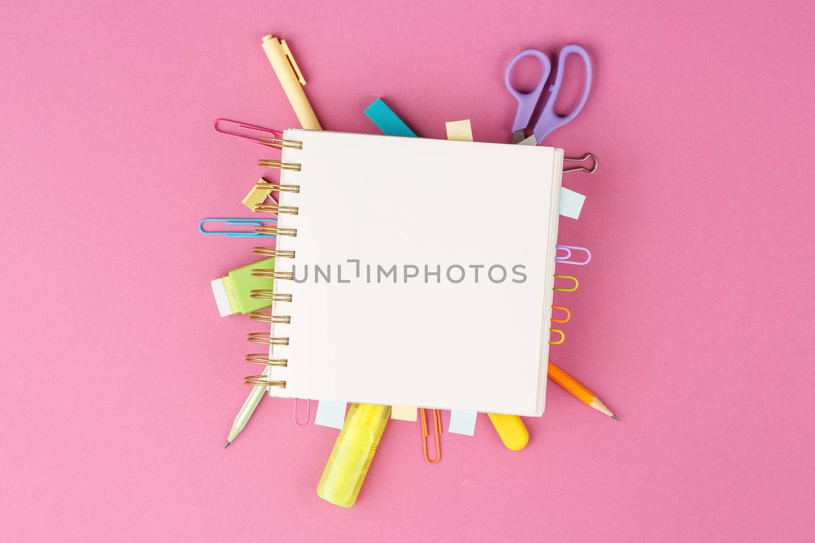 Open spiral notebook with bookmarks from paper clips and leaves for notes, pencil, scissors, binders on a pink background. Top view. Stationery concept. Office tools.