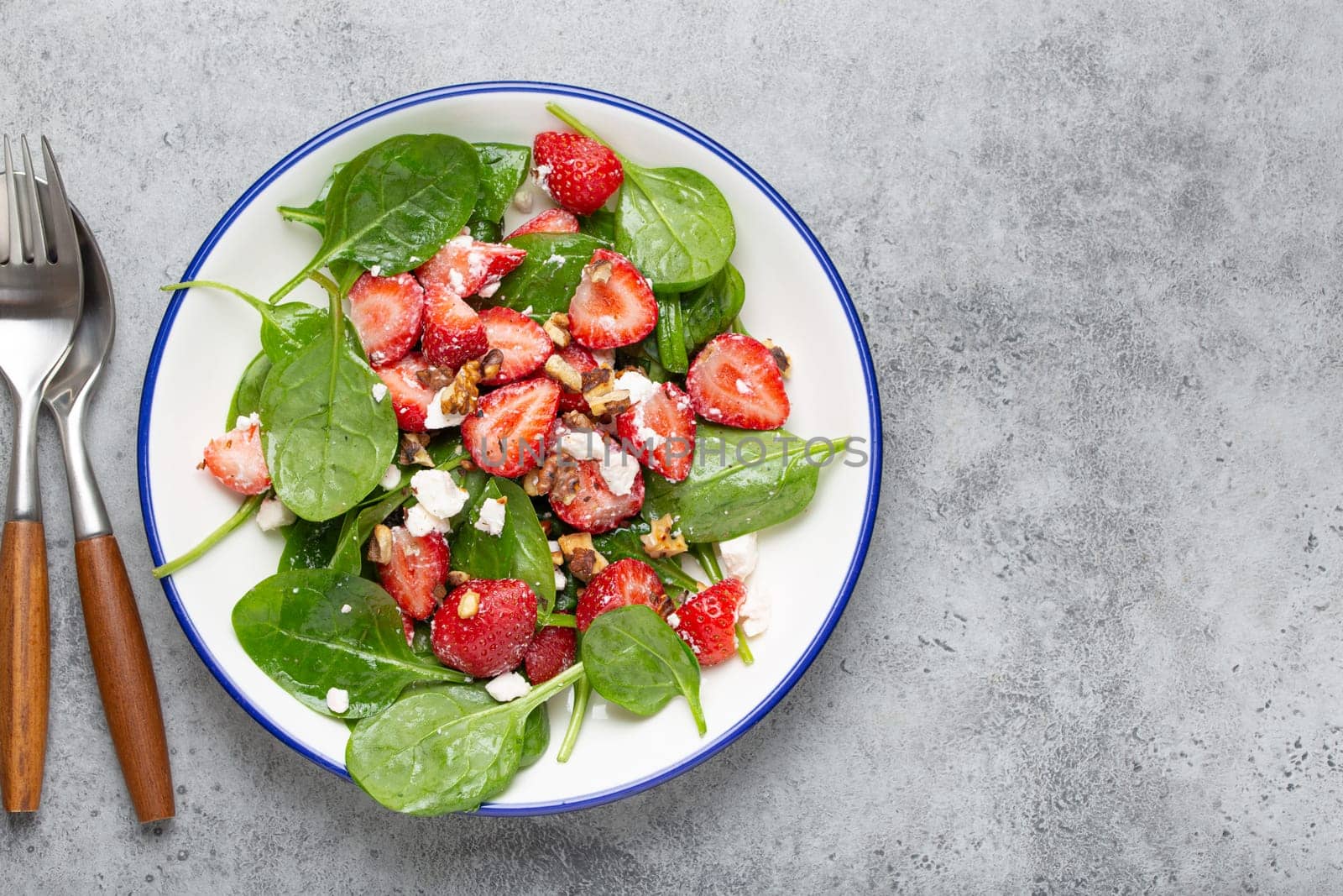Light Healthy Summer Salad with fresh Strawberries, Spinach, Cream Cheese and Walnuts on White Ceramic Plate, Grey rustic stone Background Top View Copy Space.