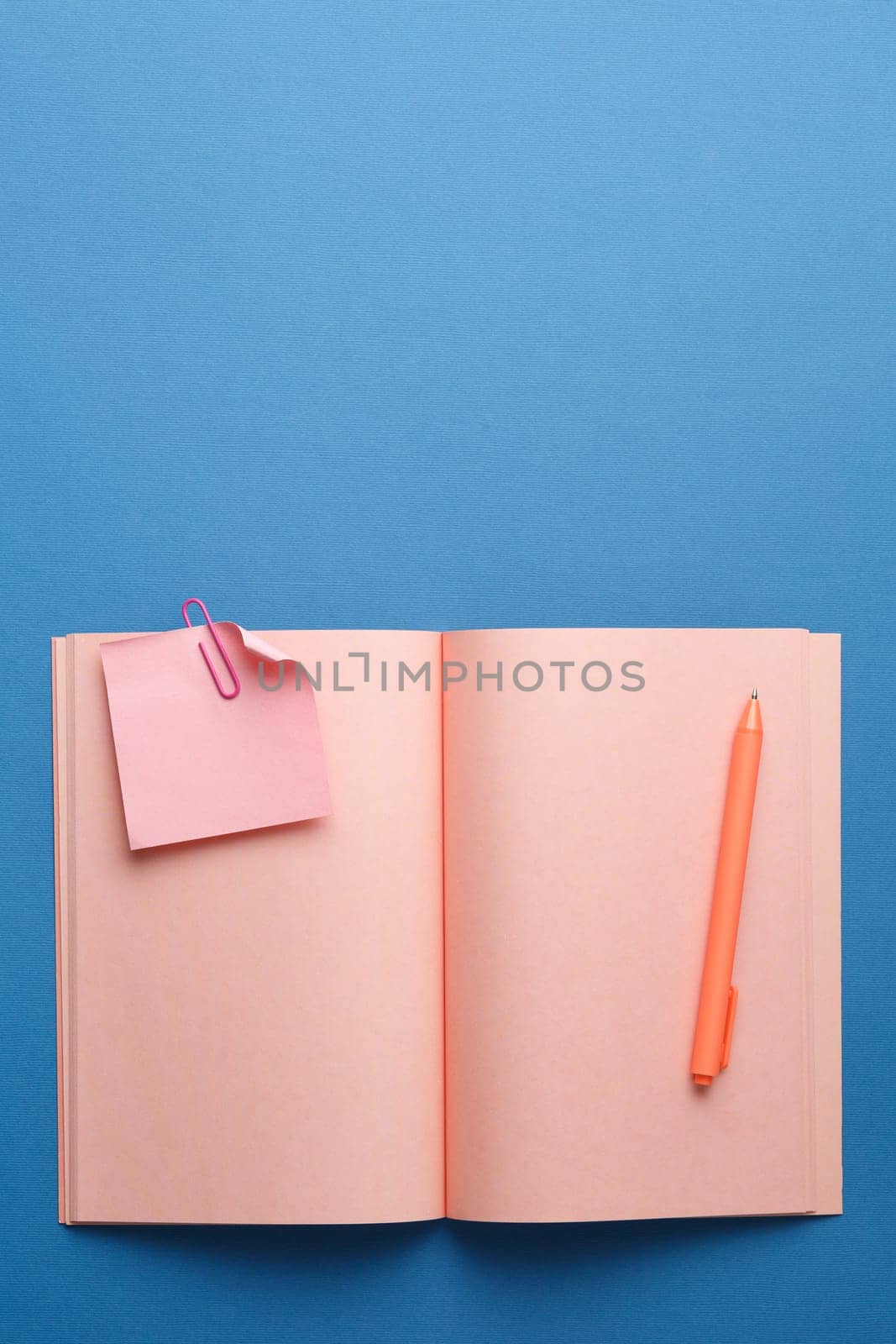 Pink notebook with pen, note sheet and color paper clip on blue isolated background. Flat lay. Top view. Vertical photo.