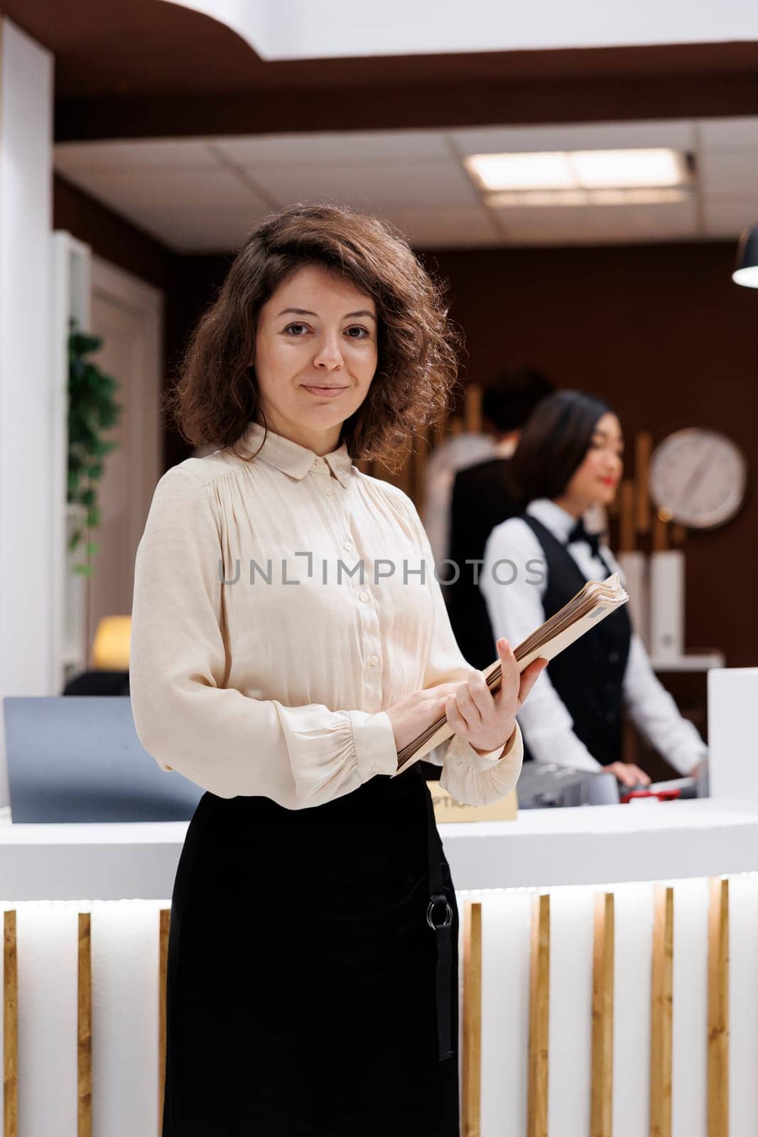Administrator carrying record files and standing near front desk in hotel lobby, verifying service as manager. Female supervisor checking booking register and welcoming people.