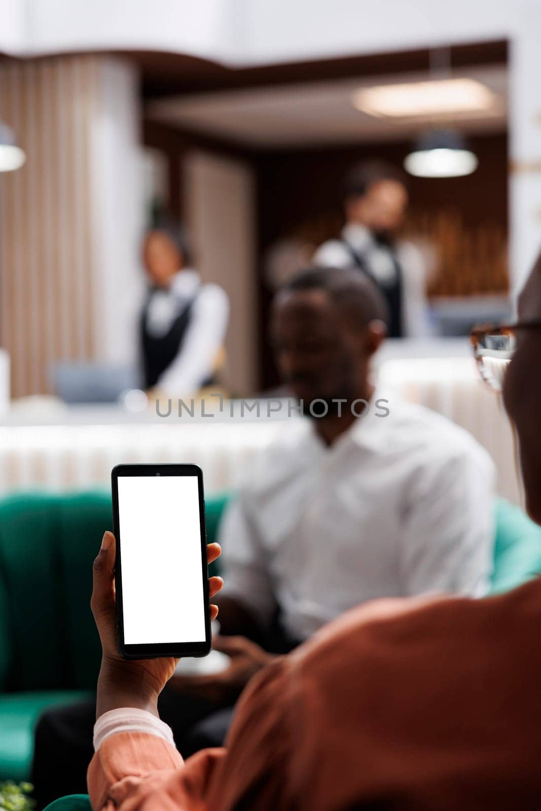 Young adult using white screen on phone, holding blank copyspace display in lounge area at hotel. Woman in resort lobby looking at isolated chromakey template, reception front desk.