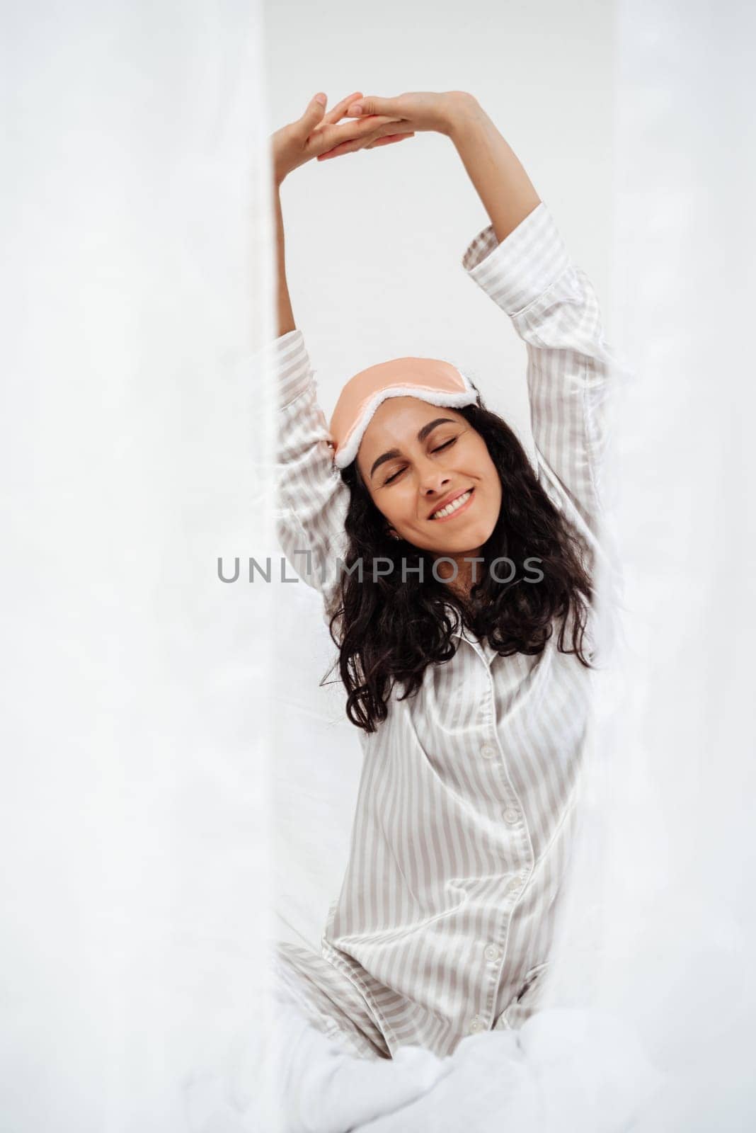 Girl sitting on bed in morning, dressed in white clothes, pulling arms up and right to wake up, stretching muscles before busy working day. Young woman of oriental appearance takes care of her health