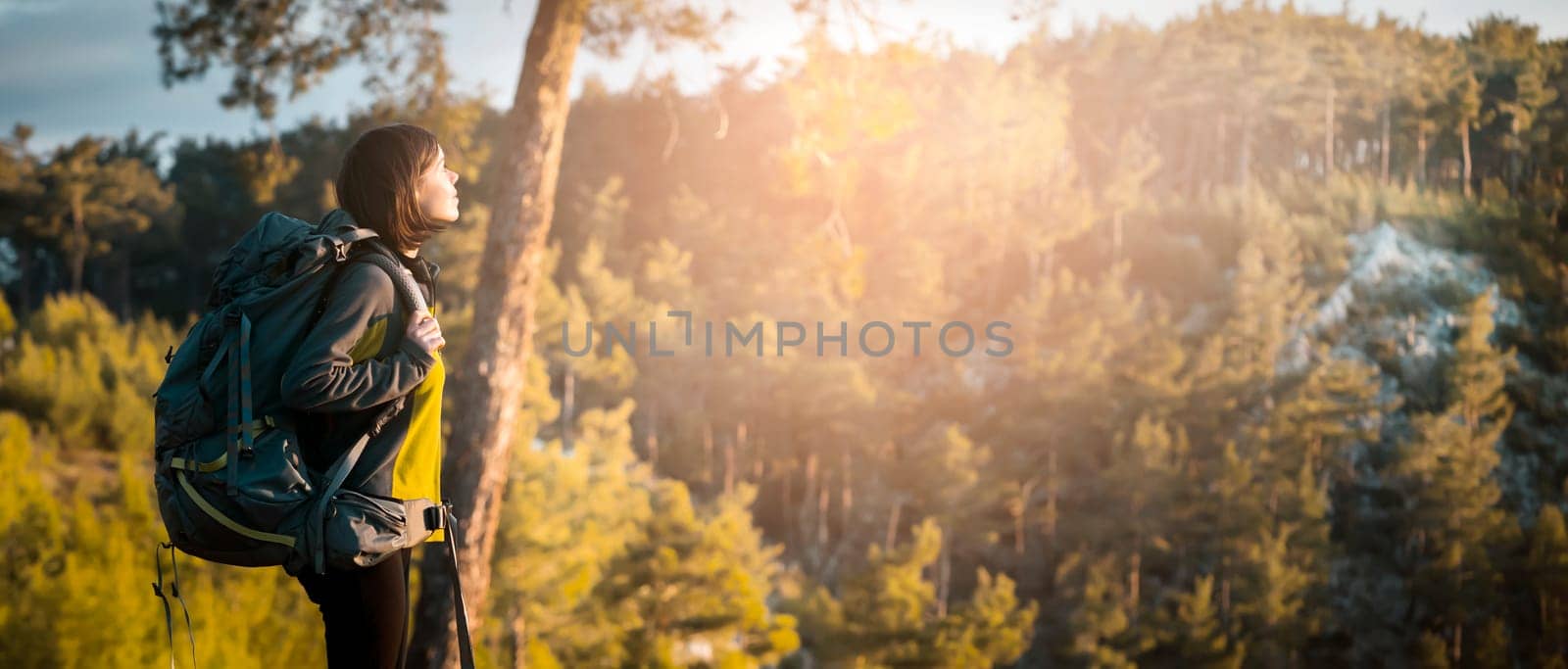 A young girl with a backpack and travel equipment looks at the amazing sunset in the mountains while she hikes along the trail. A traveler walks through the national park.