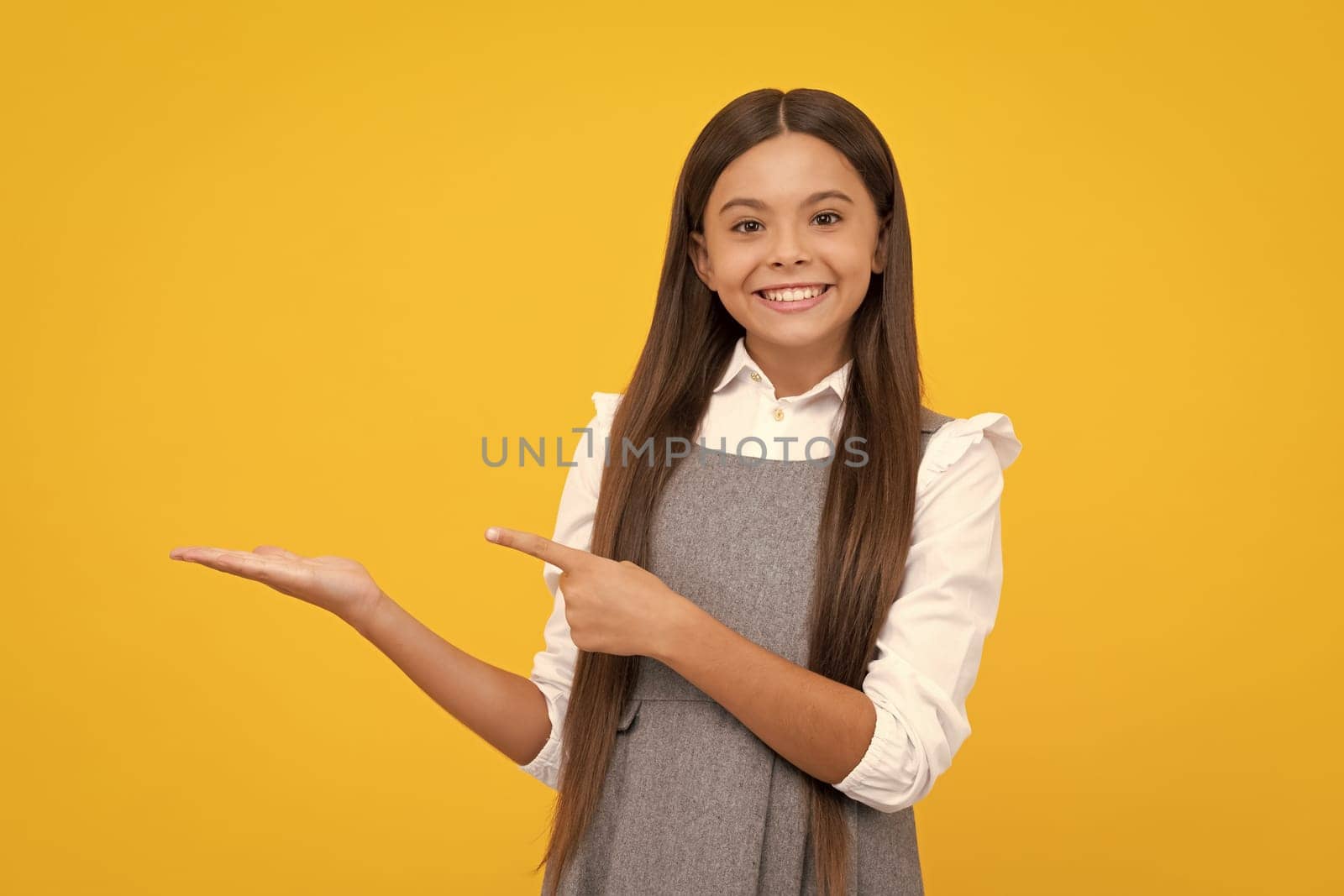 Happy girl face. Close up portrait of teenager child girl showing at copy space, pointing to ads advertising, isolated over yellow background. Mock up copy space