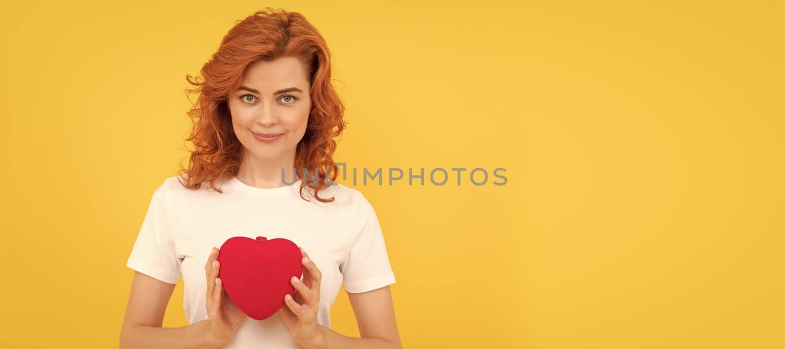 smiling redhead woman hold love heart on yellow background, 14 february. Woman isolated face portrait, banner with copy space