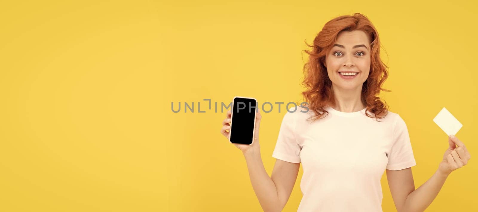 excited young lady isolated overyellow background using mobile phone holding credit card. Woman isolated face portrait, banner with mock up copy space. by RedFoxStudio